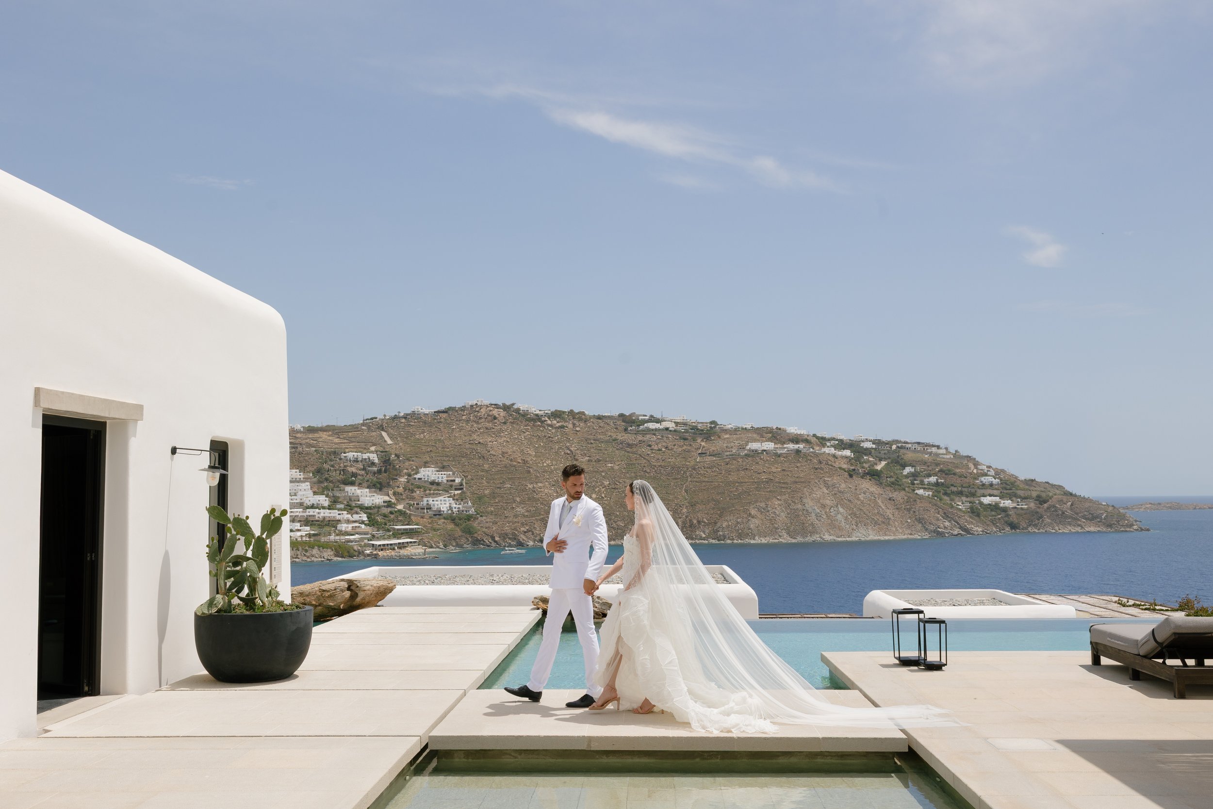 A bride and groom holding hands during their wedding ceremony outdoors by a swimming pool with ocean and hillside background, white modern building on the left, potted plant, and scenic view under a blue sky.