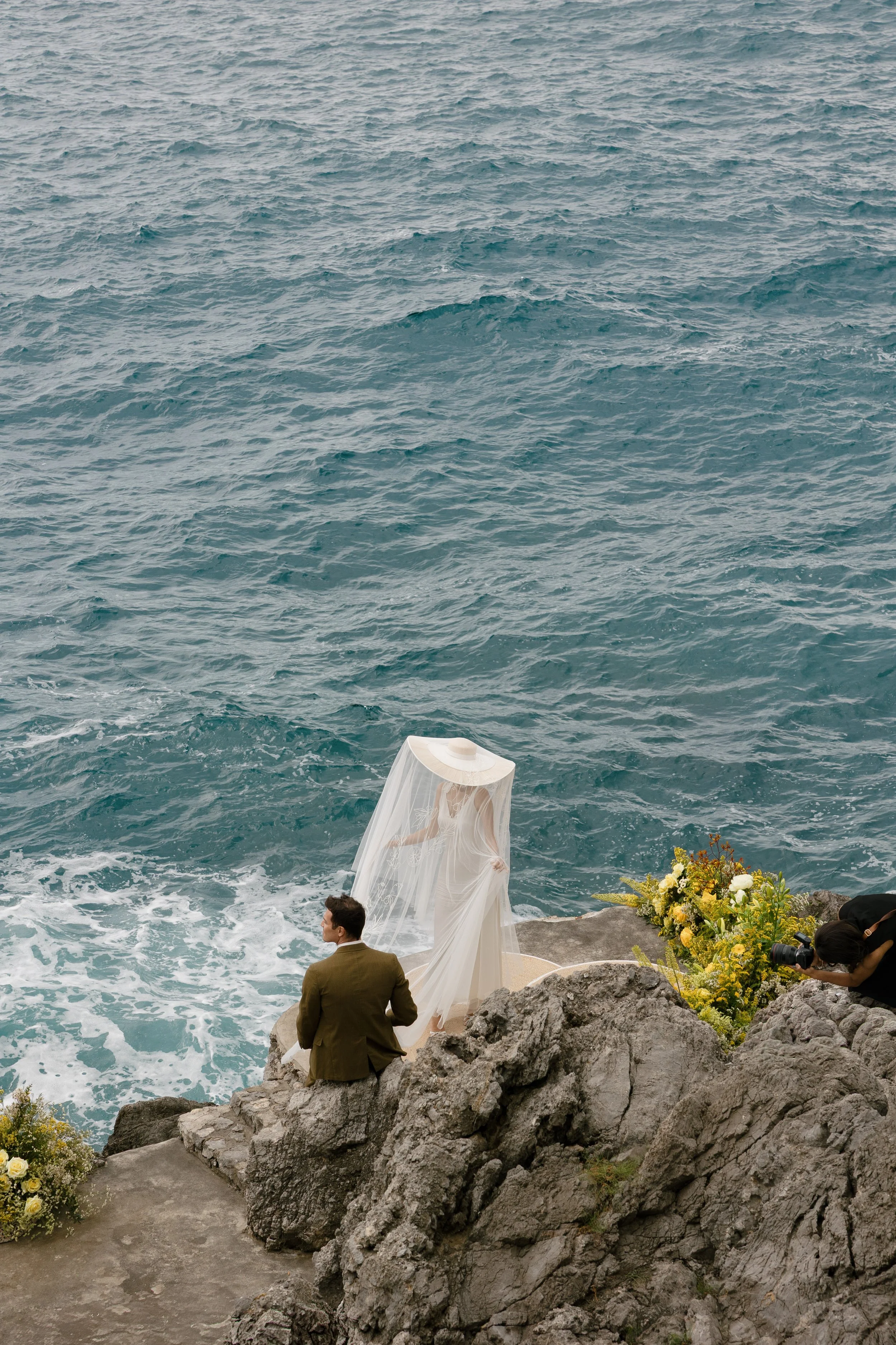 A couple's wedding ceremony on a rocky shoreline with ocean waves, flowers, a bride in a white dress with veil, groom in a suit, and a photographer capturing the moment.