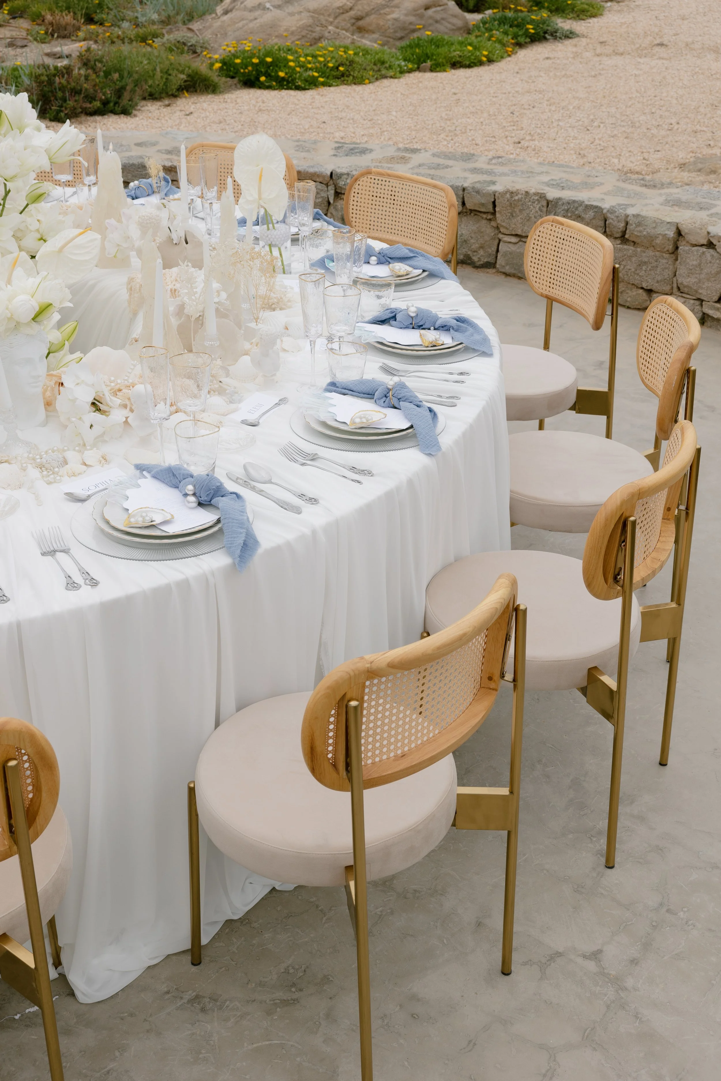Elegant outdoor dining table set with white tablecloth, floral centerpiece, candles, and place settings with blue napkins, glasses, and silverware. Surrounding beige chairs with wooden frames.