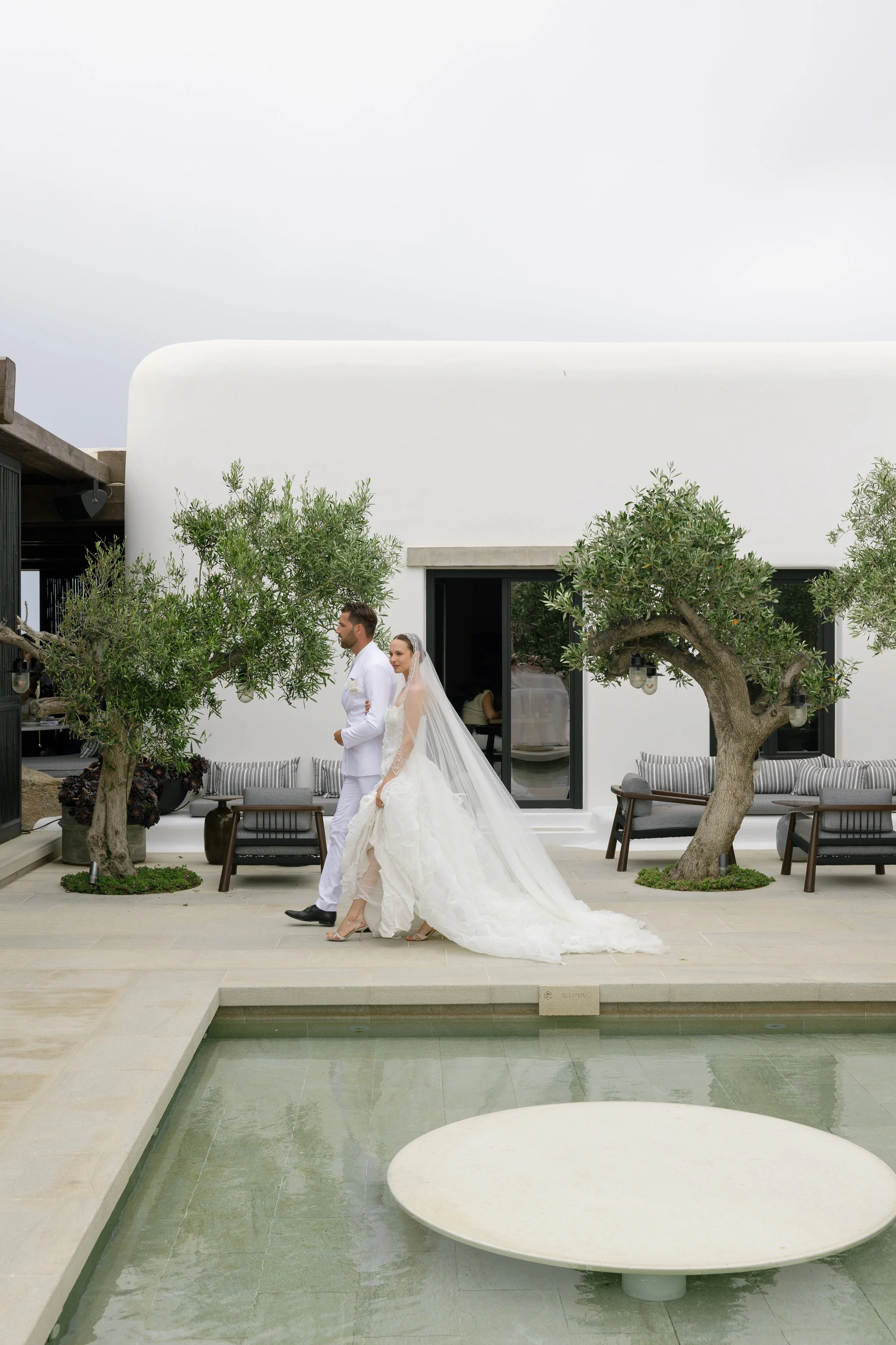 A bride in a white wedding gown and veil walking with a groom in white suit outside in a modern courtyard with trees and outdoor furniture.