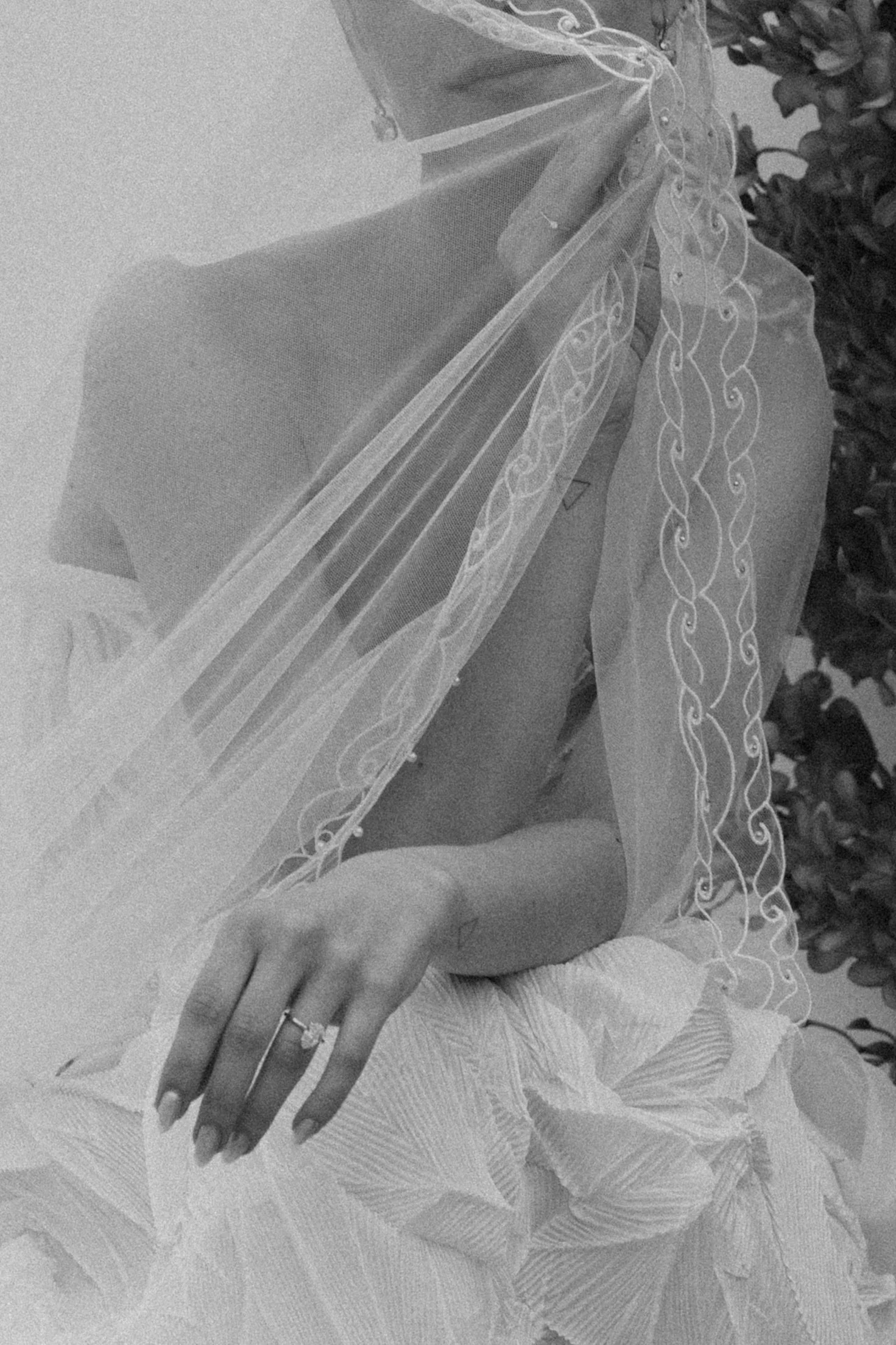 Close-up of a person's hand with rings on the fingers, resting on a textured fabric surface. The person is wearing a lace garment with intricate embroidery and tulle fabric.