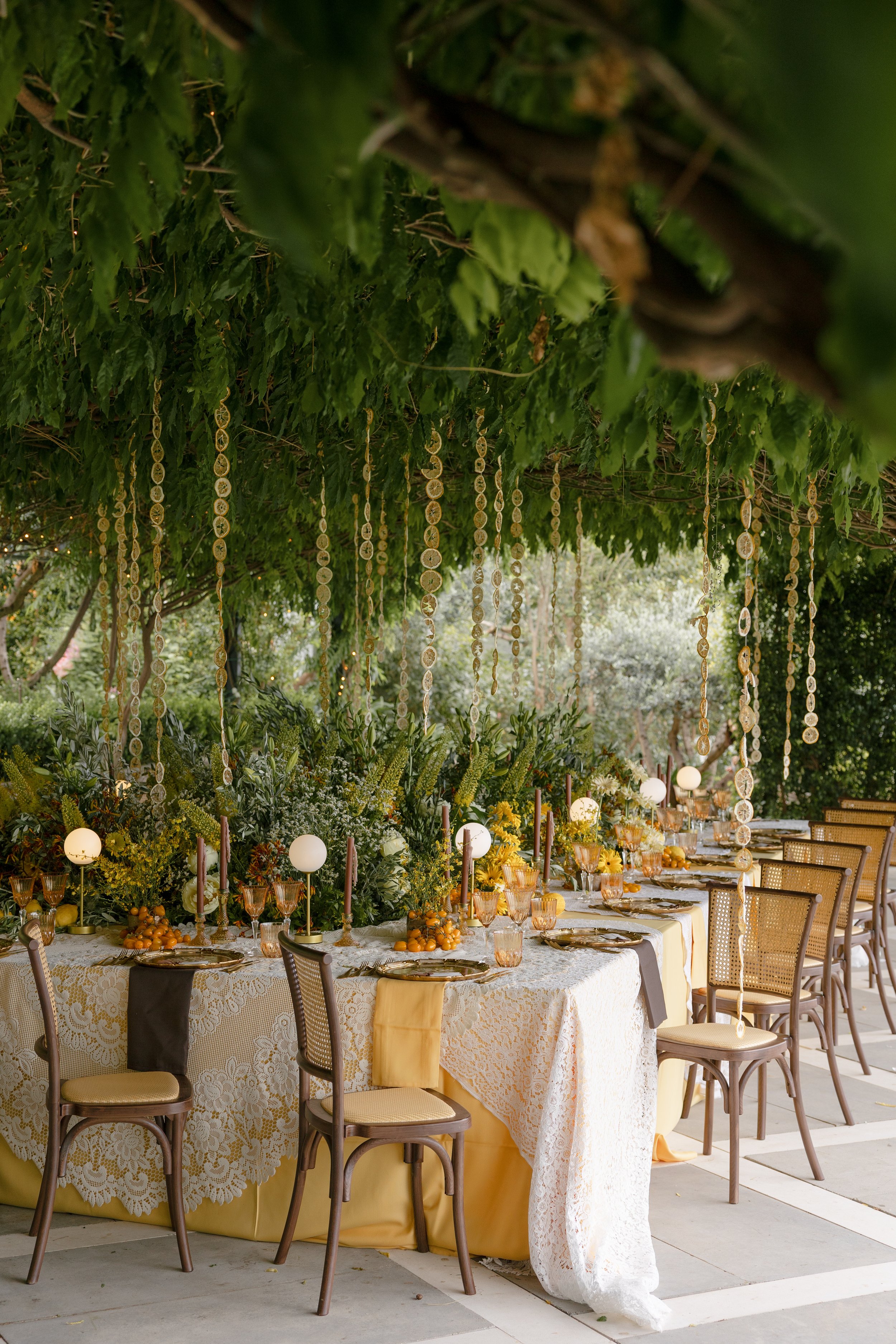 A long banquet table decorated for a celebration, with a yellow and white lace tablecloth, gold and amber glassware, floral arrangements, and hanging lemon slices from above, under a canopy of green leaves and vines.