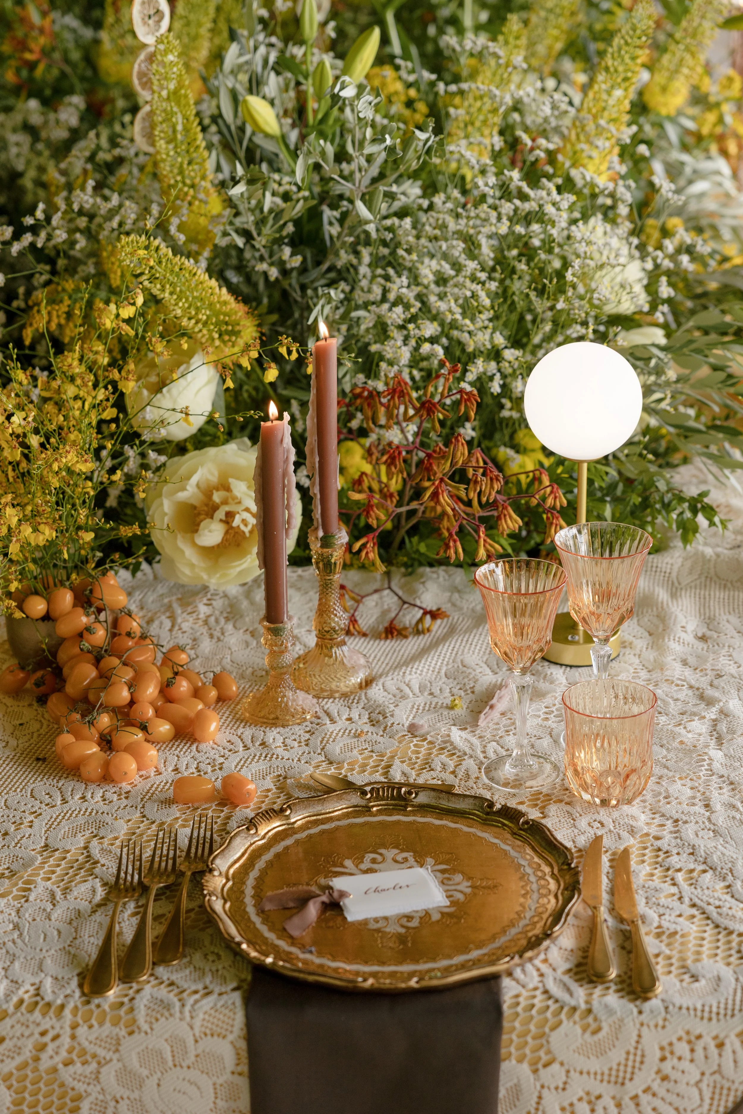 Elegant table setting with a gold-rimmed plate, gold utensils, and a lace tablecloth. The centerpiece includes white and yellow flowers, candles, and a white globe lamp. There are peach-colored glasses and a small pile of orange jelly beans on the ta
