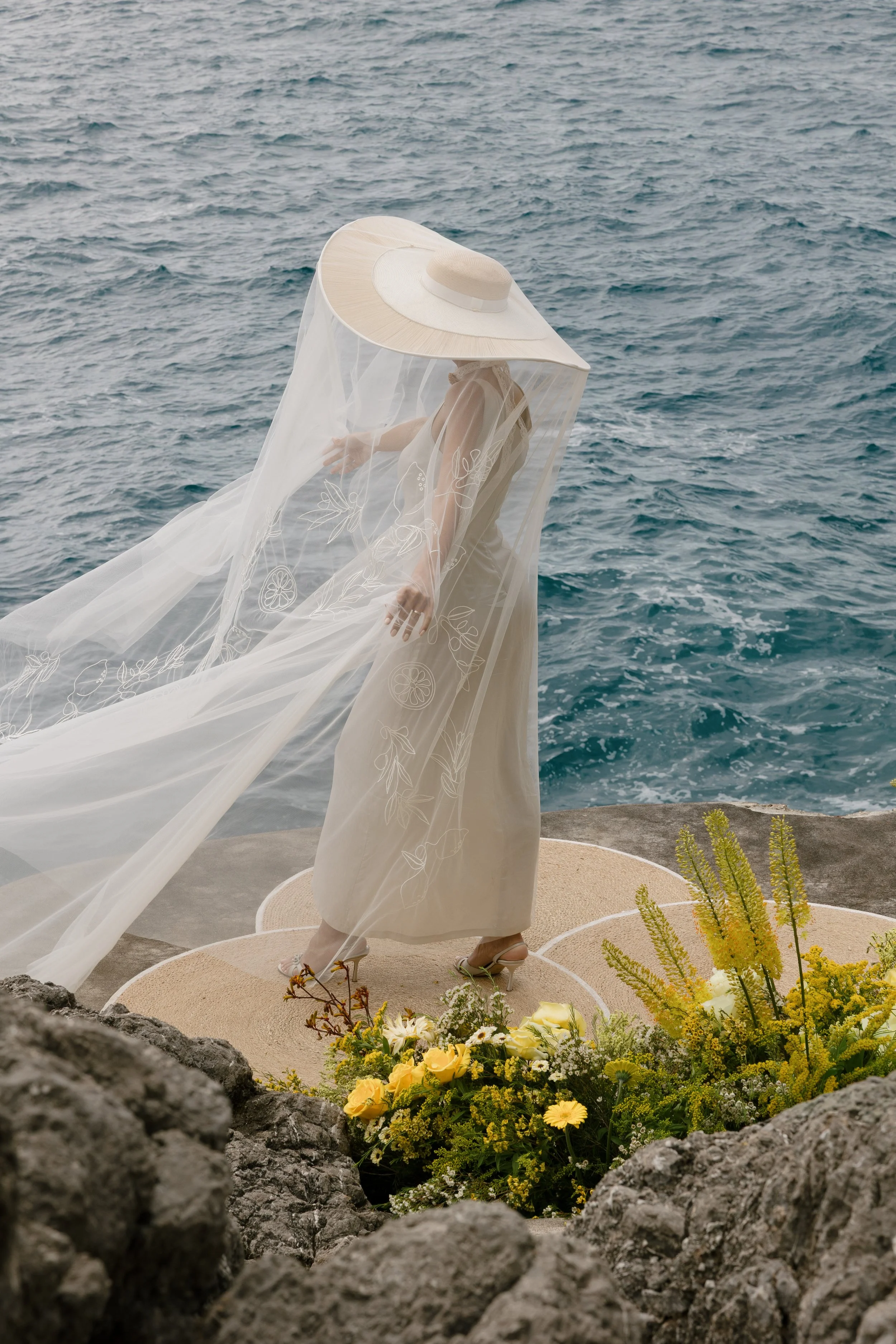 A person dressed in a beige dress, wearing high heels, standing on a circular platform surrounded by flowers, near the ocean. The person is wearing a large, wide-brimmed hat with a veil covering their face and shoulders, and the hat has floral embroi