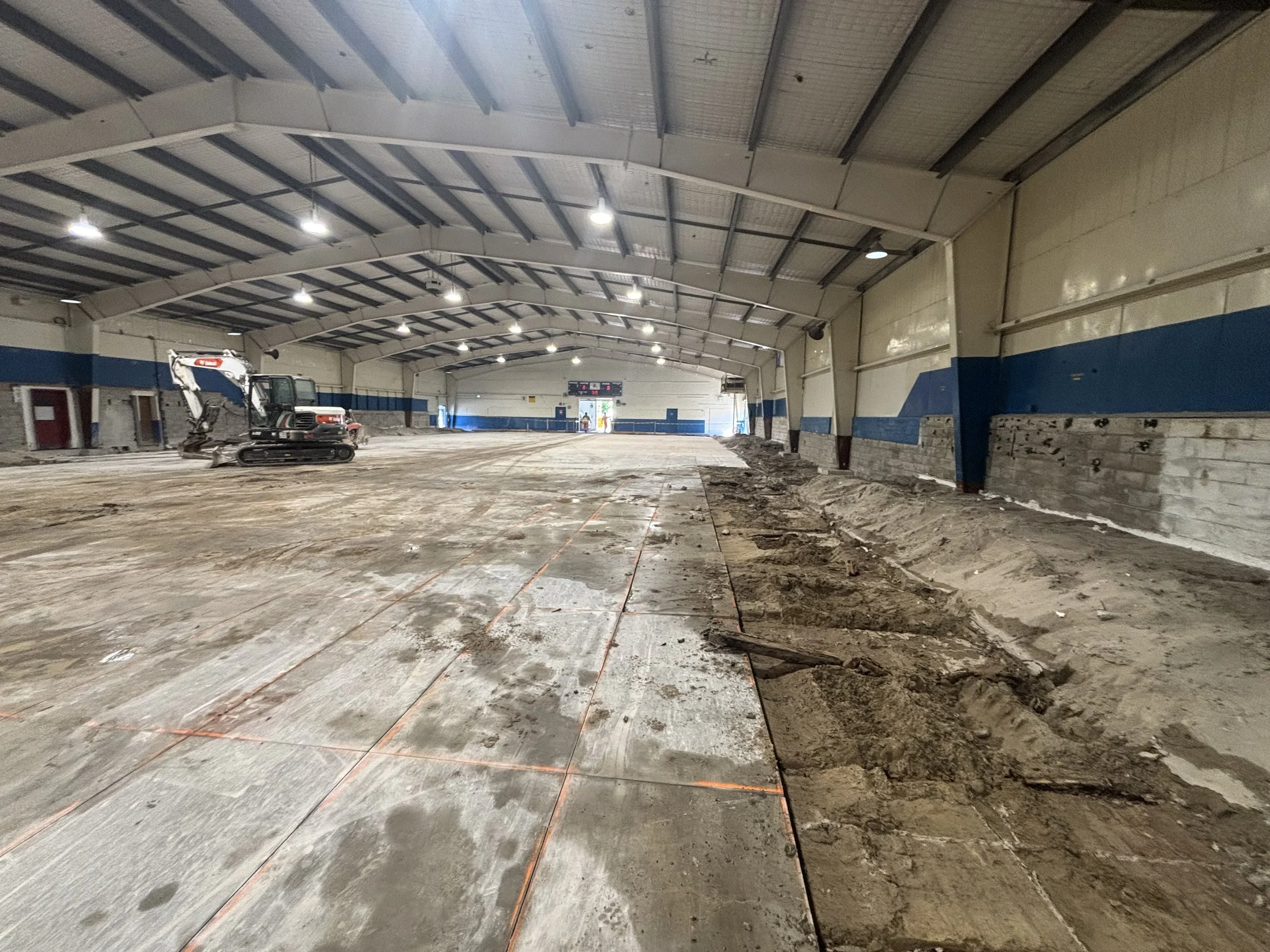 Indoor sports arena under construction with dirt and concrete floor, construction equipment, and a digital scoreboard.