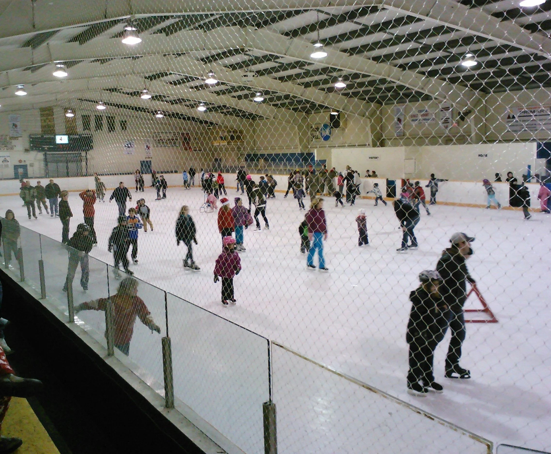 An indoor ice skating rink filled with people skating and socializing, viewed through a wire fence.
