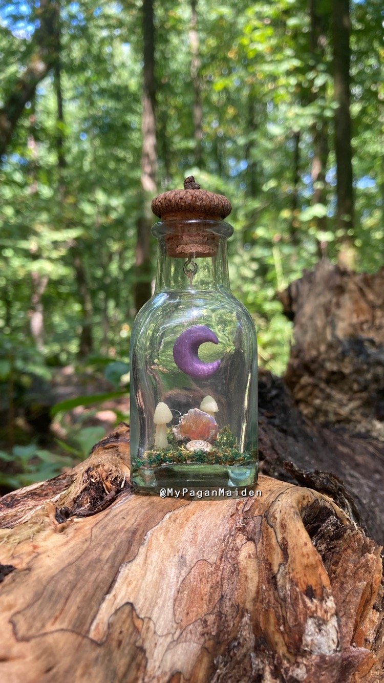 A terrarium bottle with an acorn cap lid, featuring a purple crescent moon inside along with crystals, glow in the dark mushrooms, and other nature-inspired details.