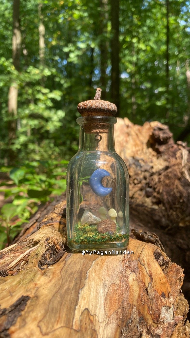 A terrarium bottle with an acorn cap lid, featuring a blue crescent moon inside along with crystals, glow in the dark mushrooms, and other nature-inspired details.