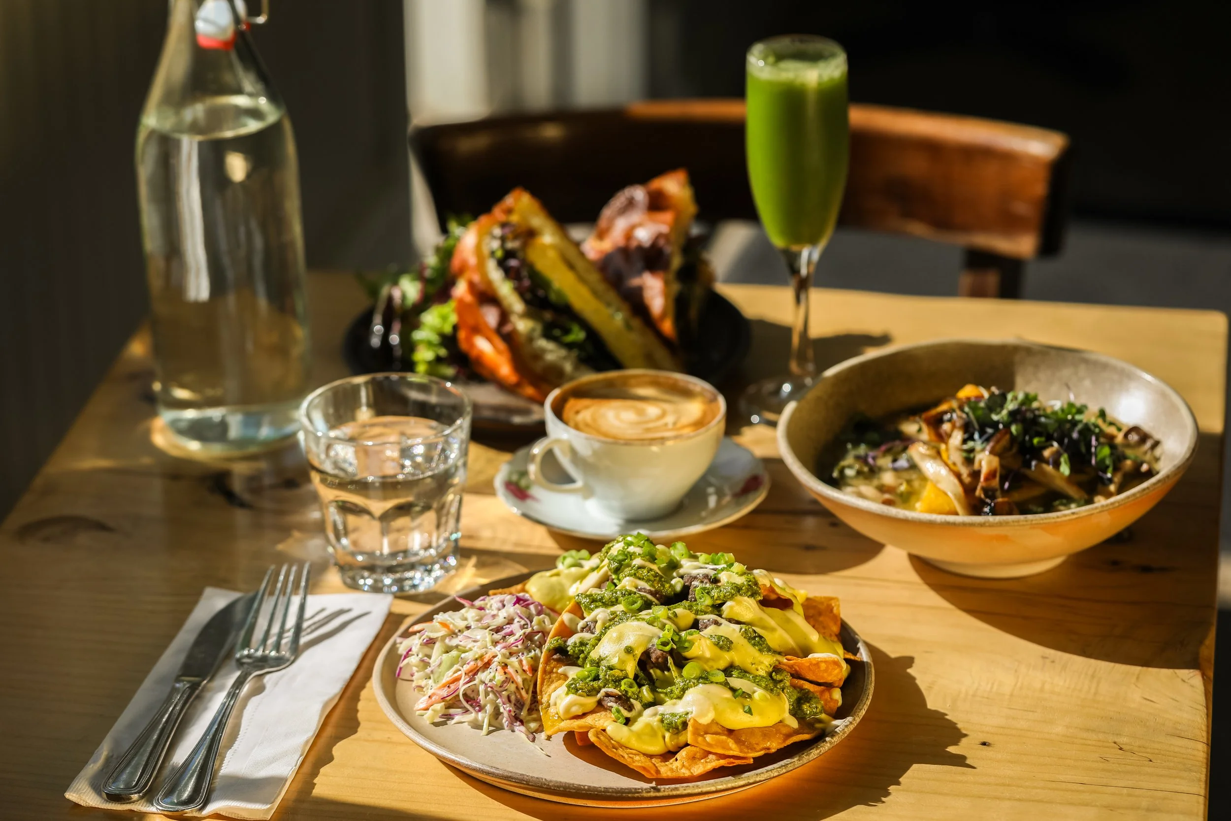 tablescape with a plate of cosmic queso nachos, a bowl of beans and greens, a latte, green juice and a frittata sandwich in the background with a glass of water, carafe, and place setting