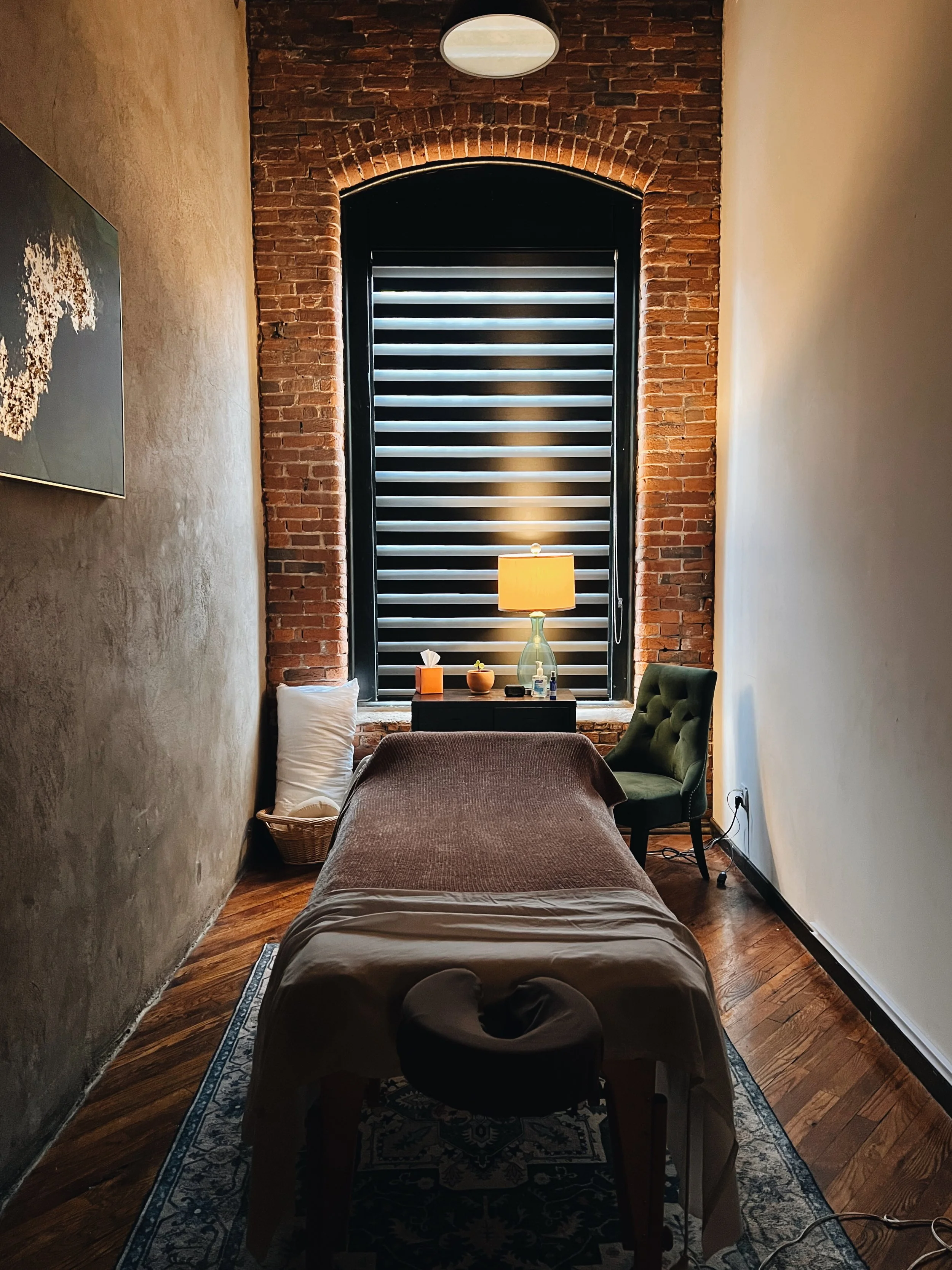 A massage room with a massage table covered by a brown blanket, a green chair to the right, a black side table with a table lamp, tissue box, and various items, a large window with black horizontal blinds, a pillow on the floor, artwork on the left wall, and hardwood floors.