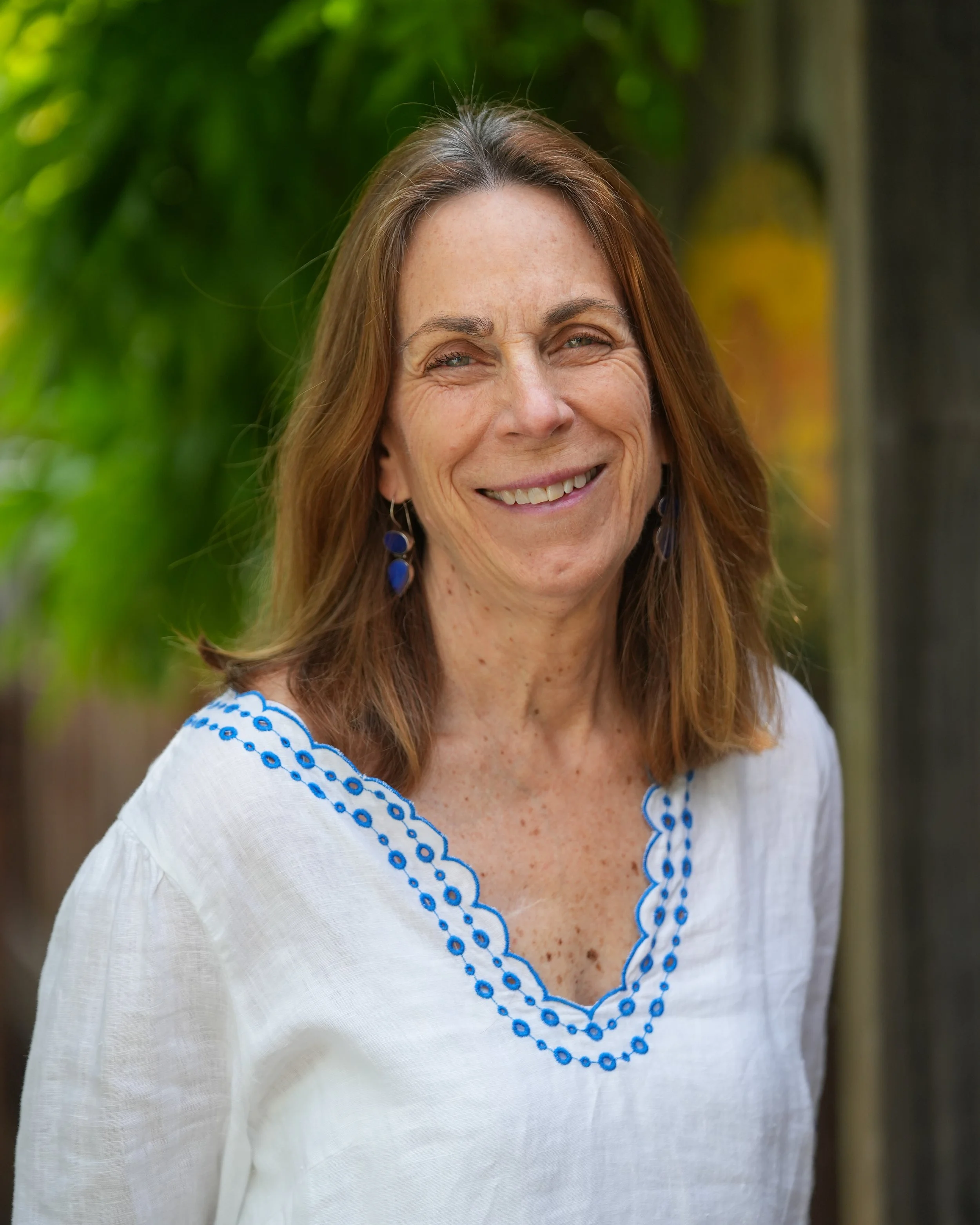 A smiling middle-aged woman with shoulder-length light brown hair, wearing a light denim jacket over a beige top, standing with arms crossed in front of a textured light blue wall.