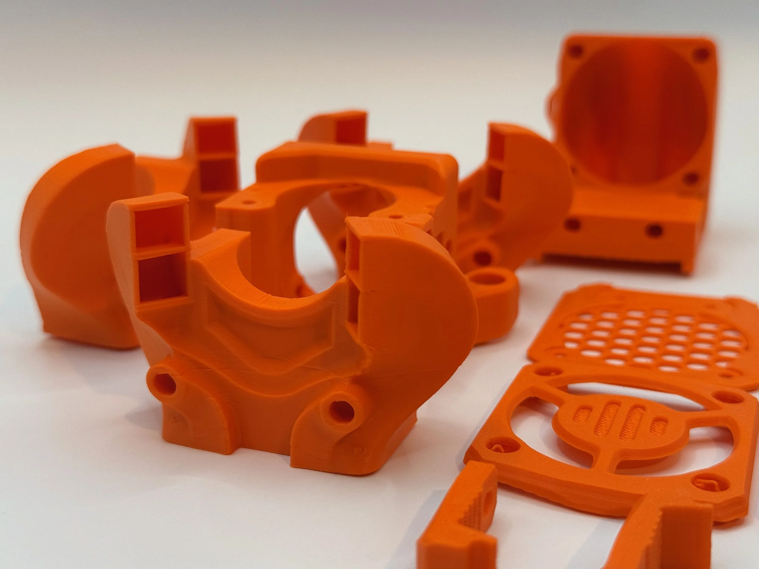 Mehrere orange 3D-gedruckte Kunststoffteile, darunter ein komplexes Gehäuse und kleinere Komponenten, auf weißem Hintergrund.