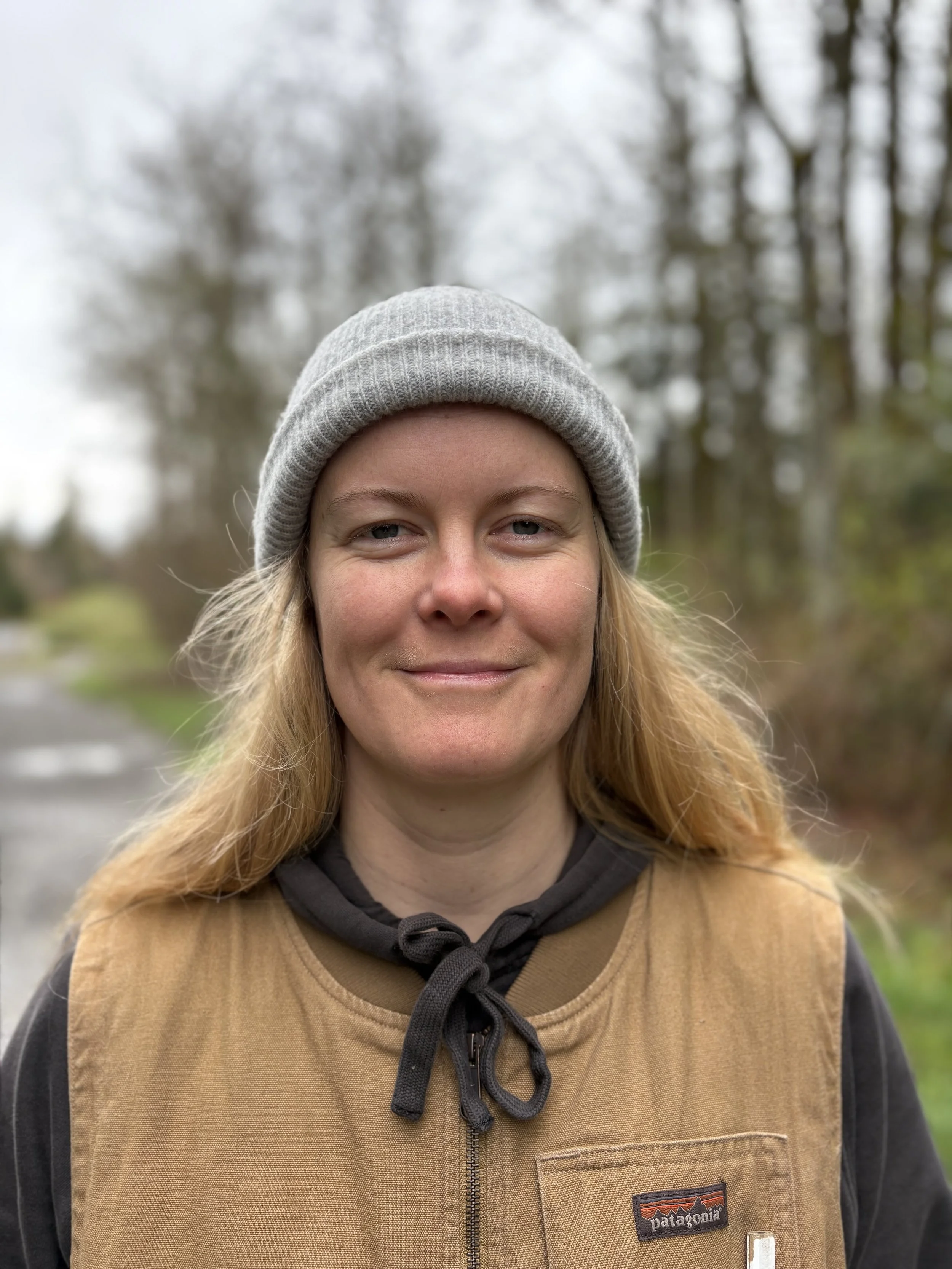 A woman wearing a gray beanie, a black hoodie with a drawstring, and a tan Patagonia vest standing outdoors among trees with a cloudy sky.
