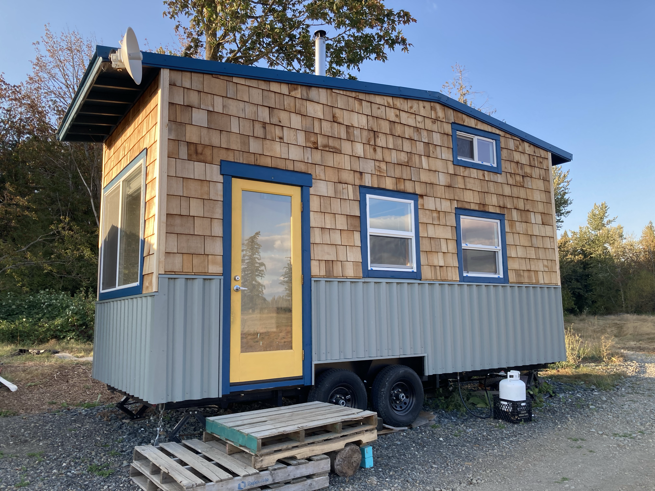 A tiny house on wheels with a yellow door and several windows, situated outdoors with trees and a clear sky in the background.