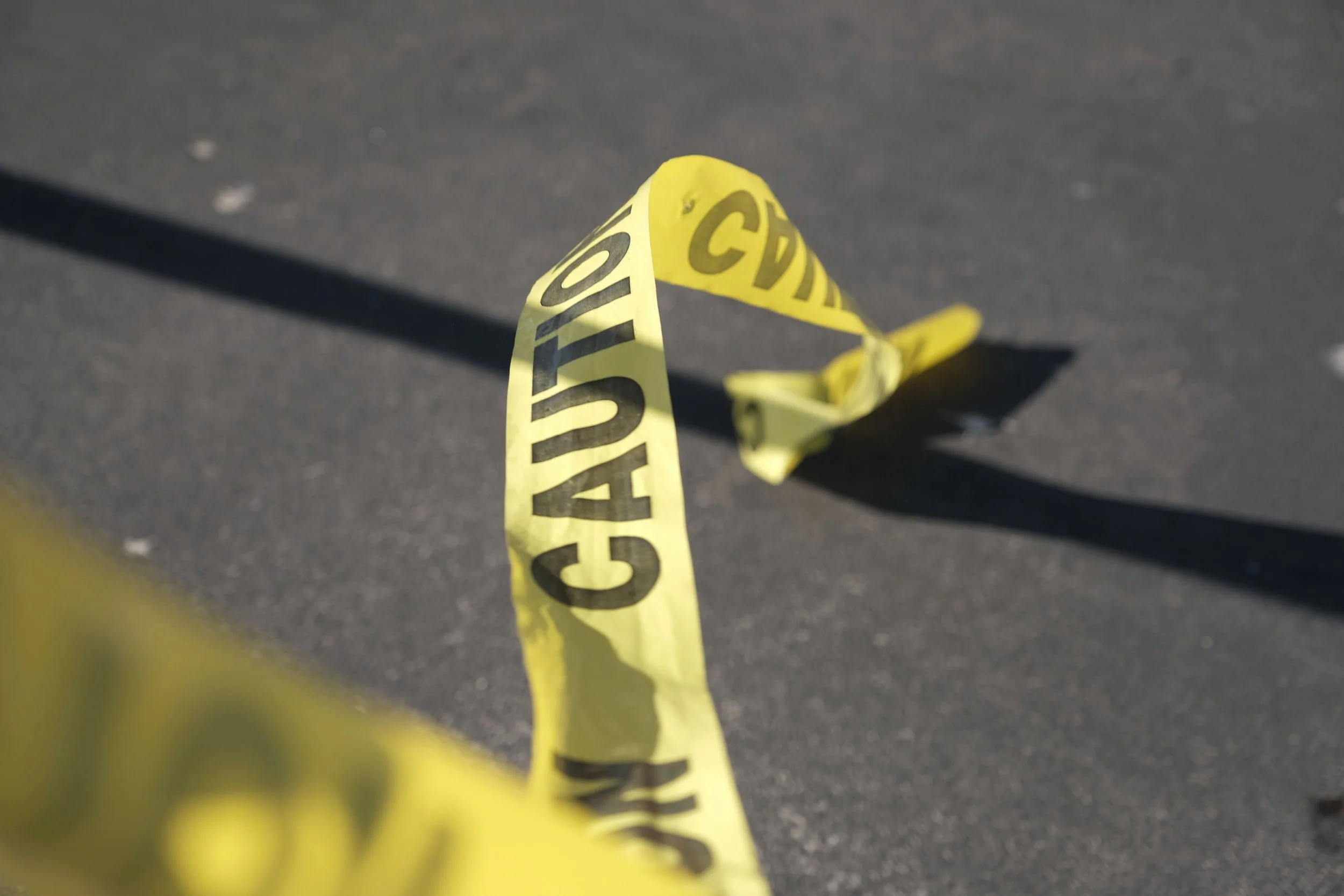 A yellow caution tape on an asphalt surface with black text, casting a shadow.