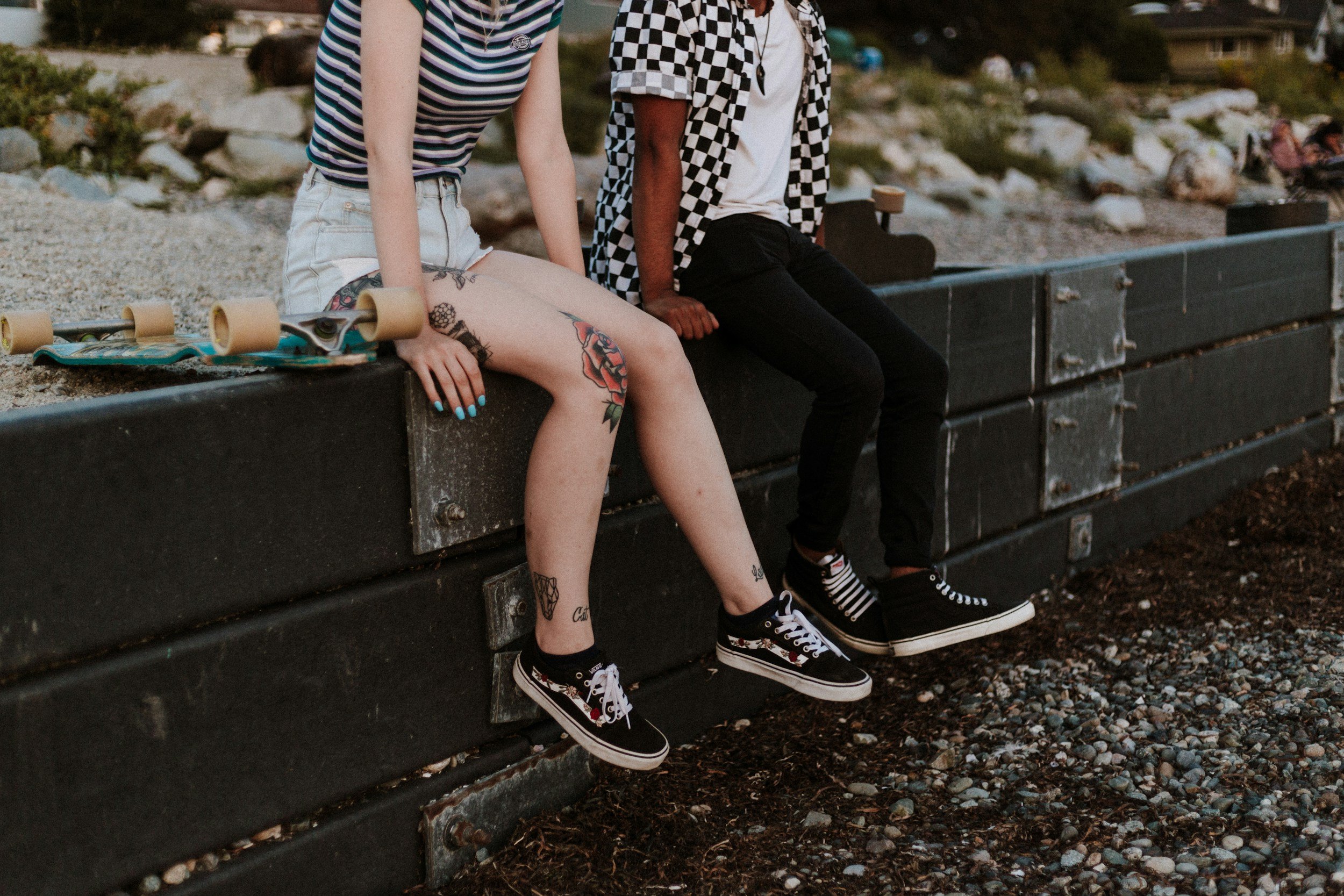 Two people sitting on a black concrete barrier at the beach. One person has tattoos on their legs and wide-legged shorts, with a skateboard beside them. The other person wears black pants, a patterned shirt, and sneakers. The background features rocks and a pebbled beach.