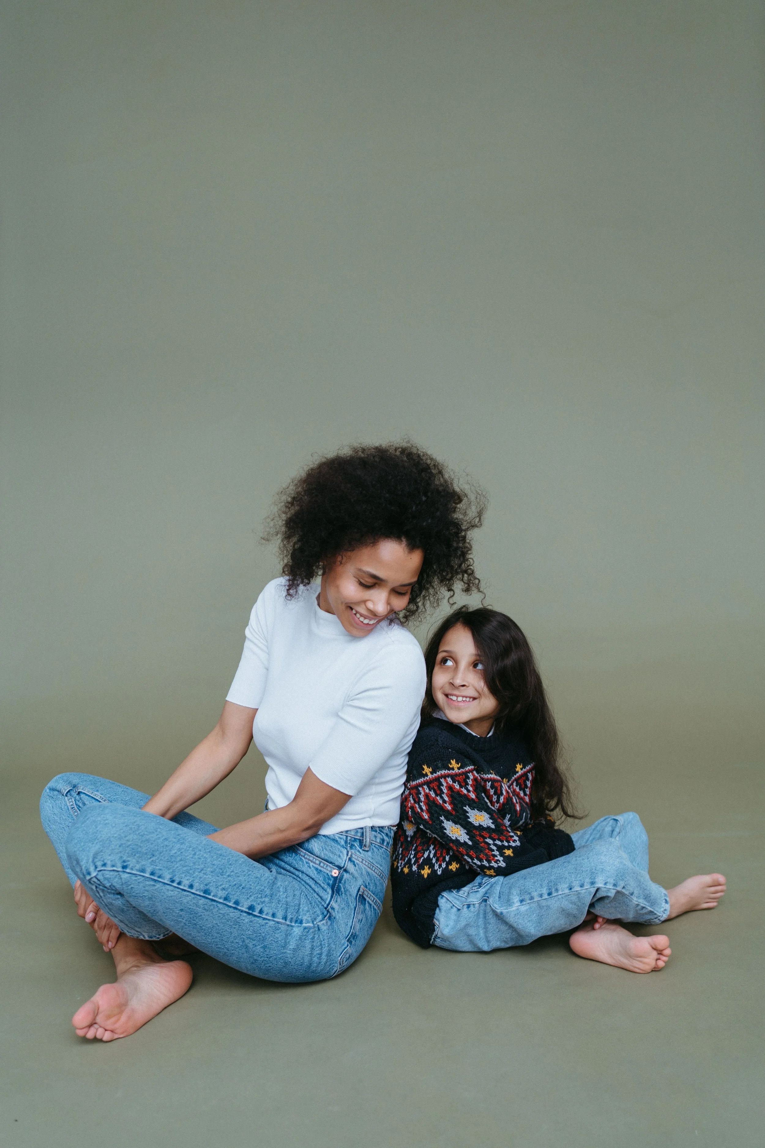 A woman with curly black hair and a young girl with long dark hair sitting on the floor against a plain green background, smiling and looking at each other. The woman wears a white shirt and blue jeans, the girl wears a black sweater with colorful patterns and blue jeans.