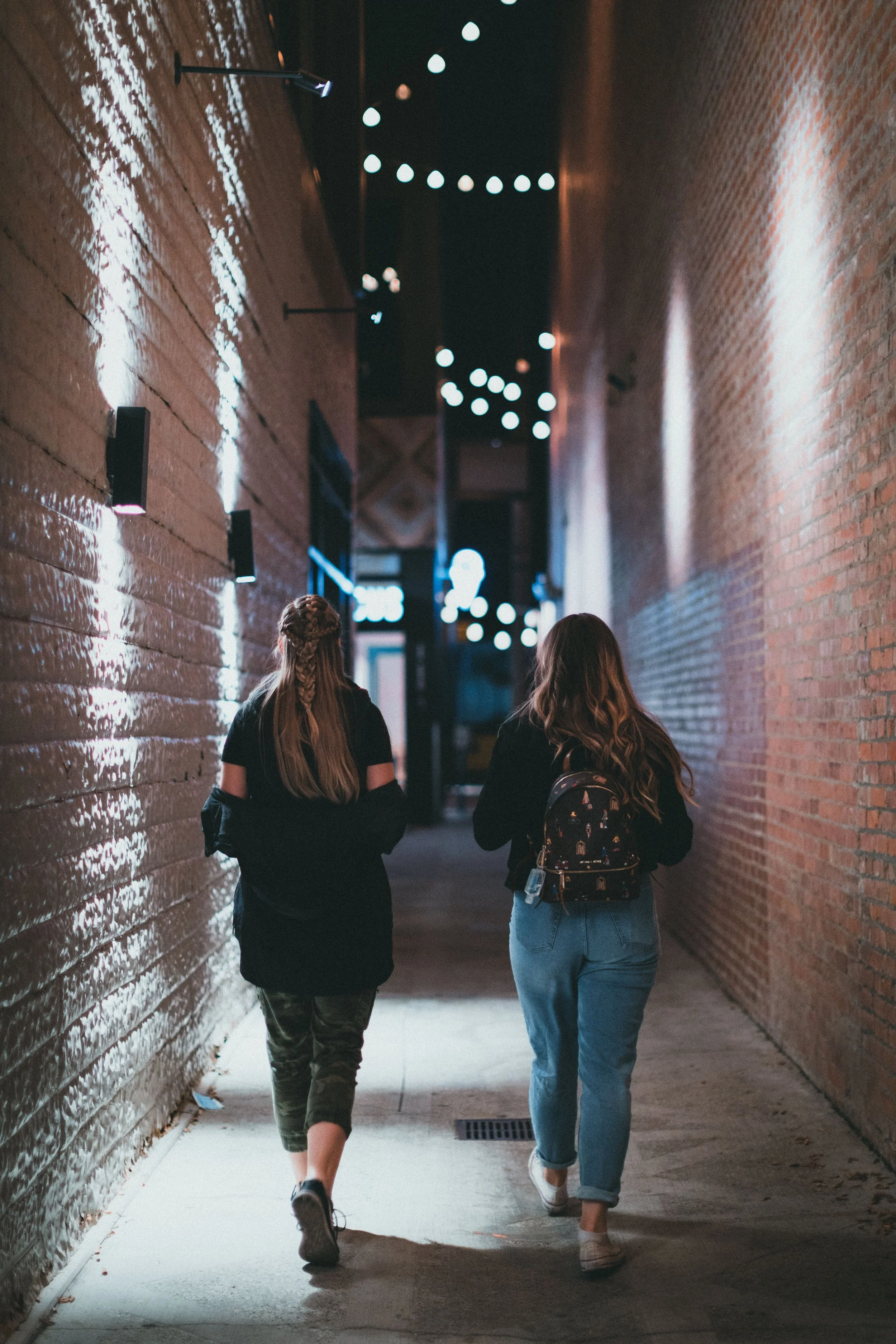 Two women walking down a narrow alley between brick buildings at night, illuminated by string lights overhead.
