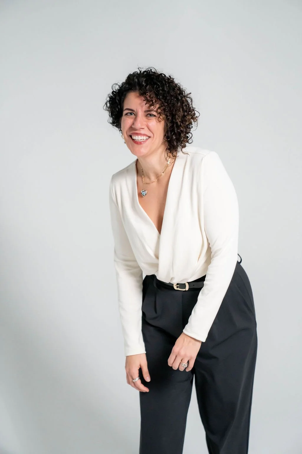 A woman with curly dark hair wearing a white blouse and black pants, smiling and leaning forward against a plain light gray background.