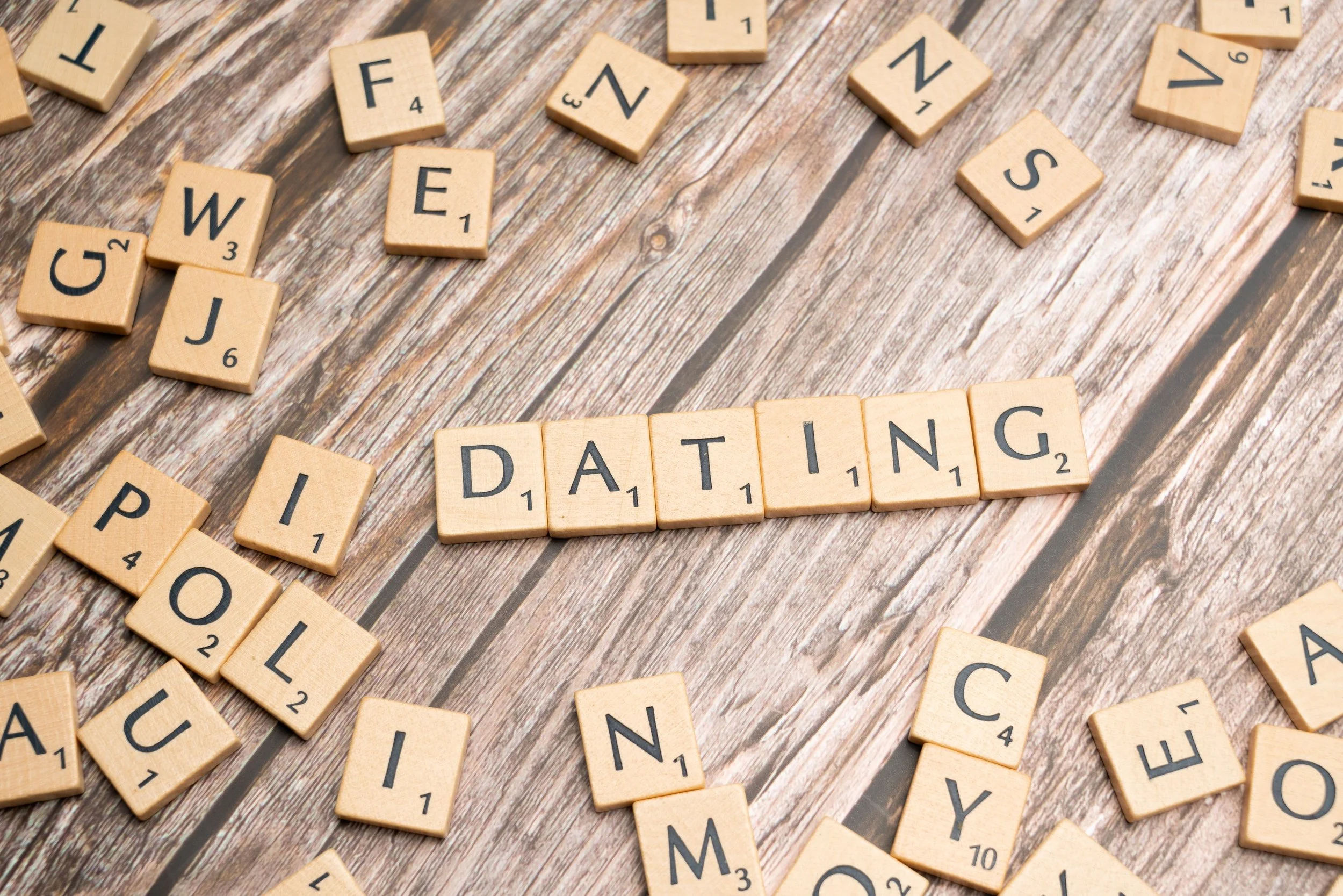 Scrabble tiles scattered on a wooden surface spelling out 'DATING' with other letter tiles around.