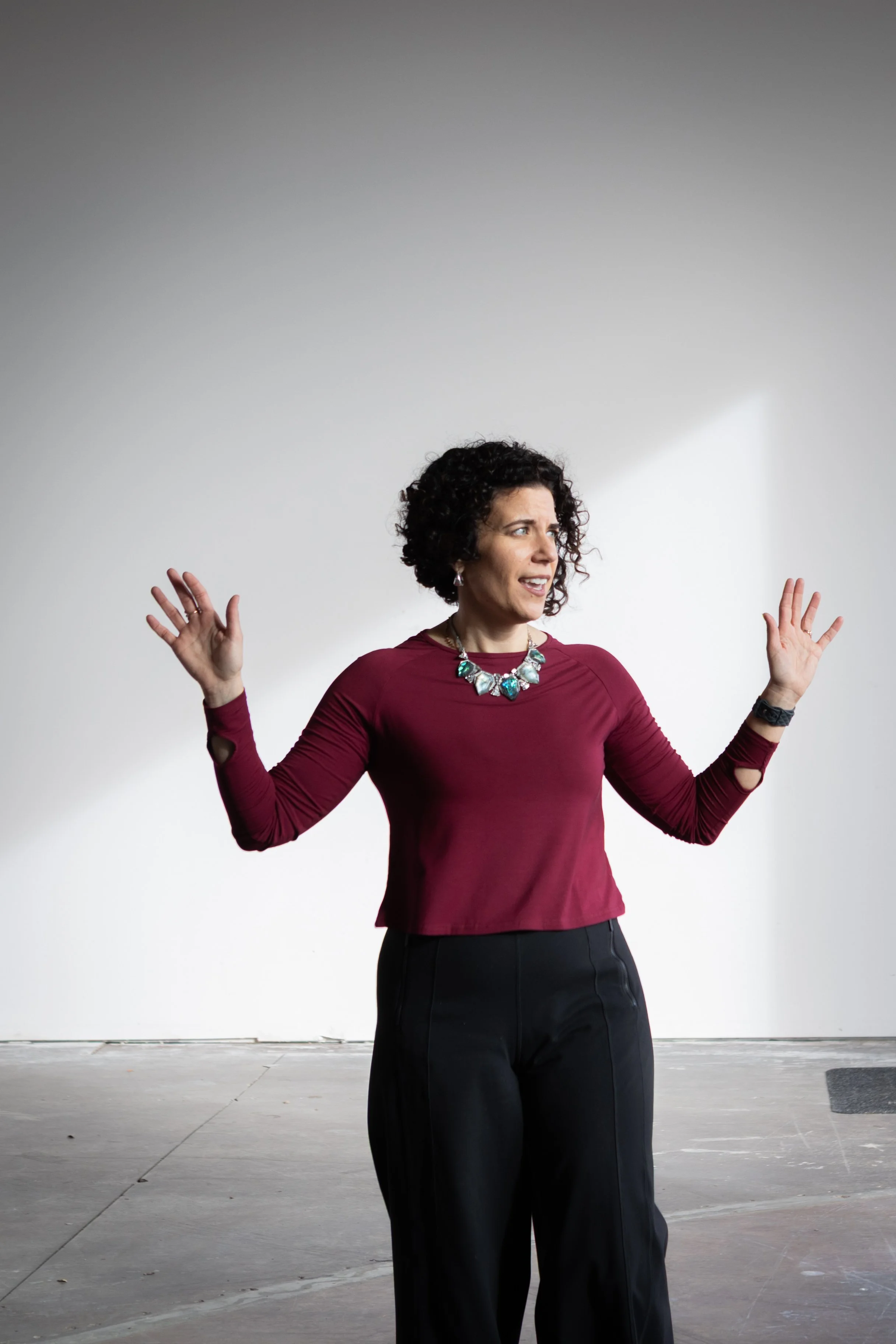 A woman with curly hair wearing a maroon long-sleeve top and black pants, gesturing with her hands and looking to the side, standing against a plain gray wall.