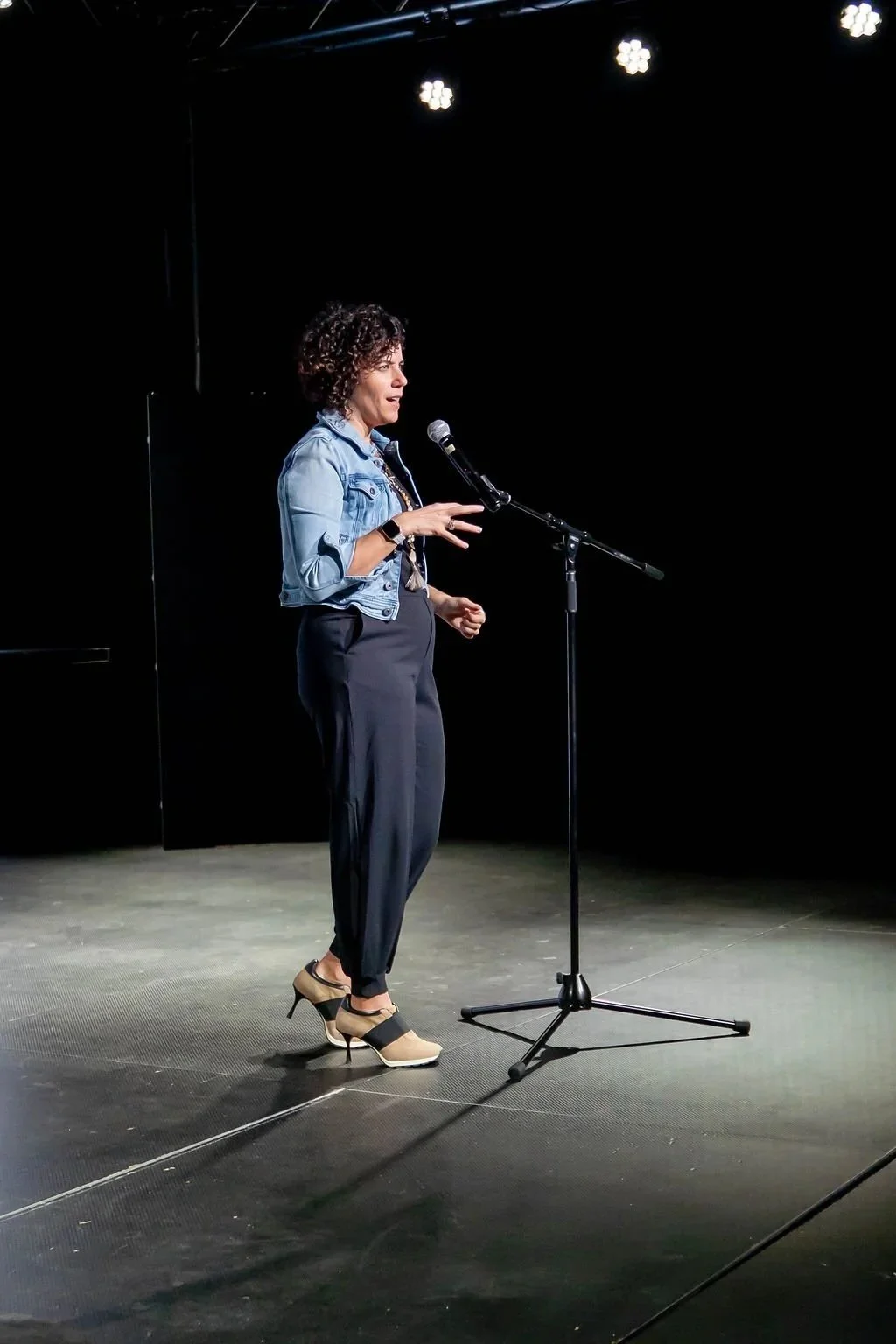A woman standing on a stage, speaking into a microphone, wearing a denim jacket, black pants, and beige shoes with black straps, against a black background with stage lights overhead.