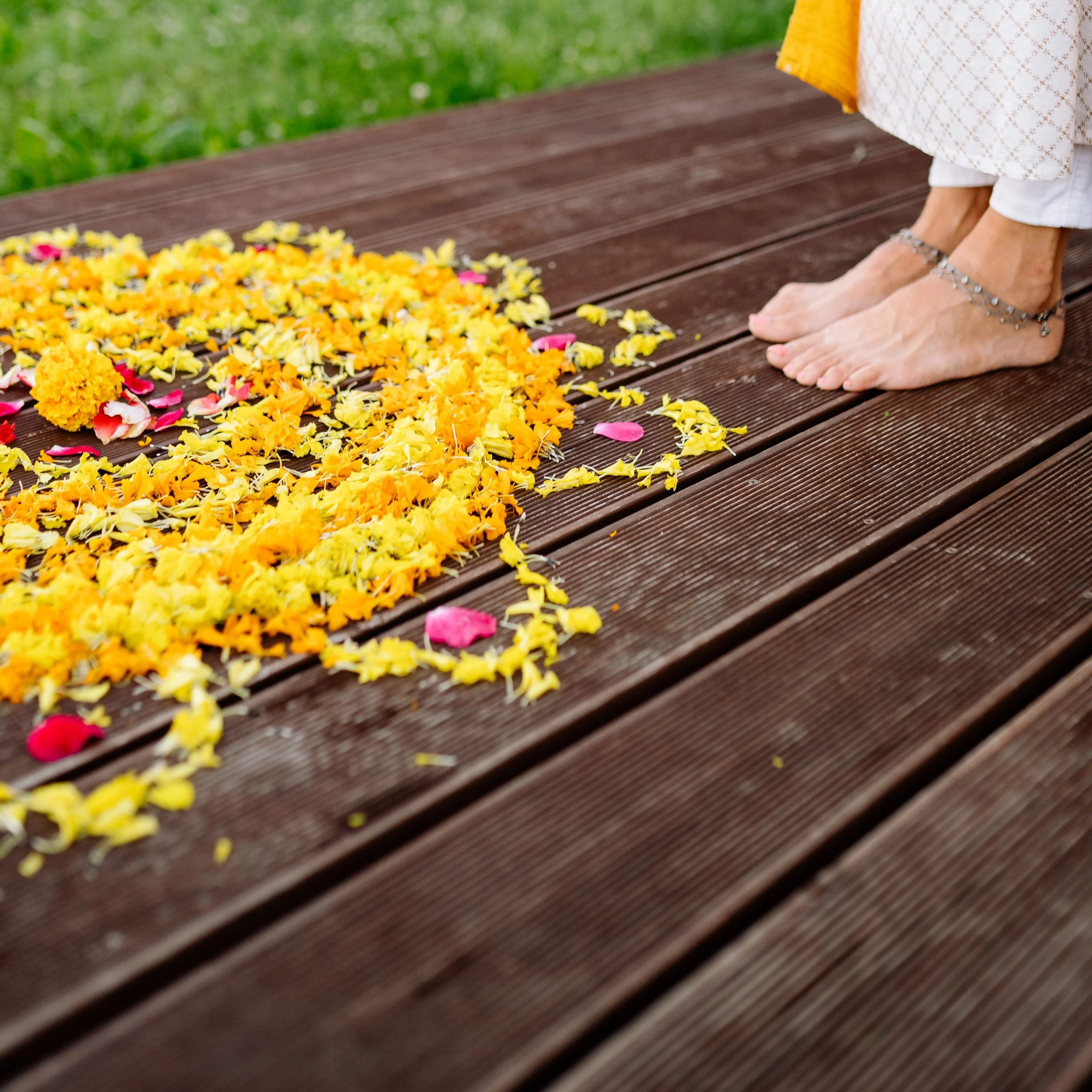 A person standing on a wooden deck with bare feet, wearing anklets and white pants, surrounded by colorful flower petals on the deck. Green garden grass is visible in the background.