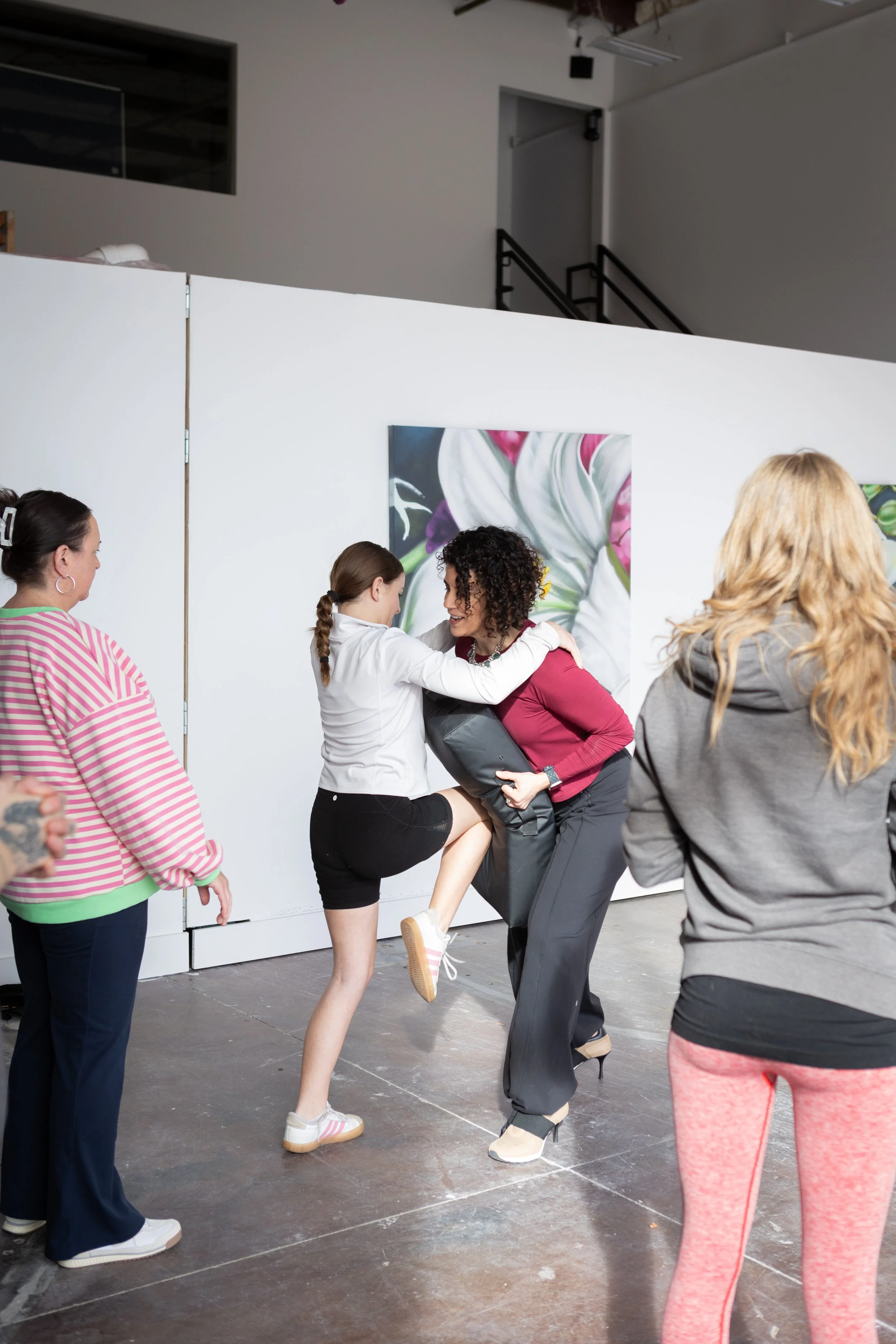 A group of women and a girl are engaged in a dance or performance in an indoor space with artwork on the wall. The girl is being lifted and supported by a woman, while others observe and participate.