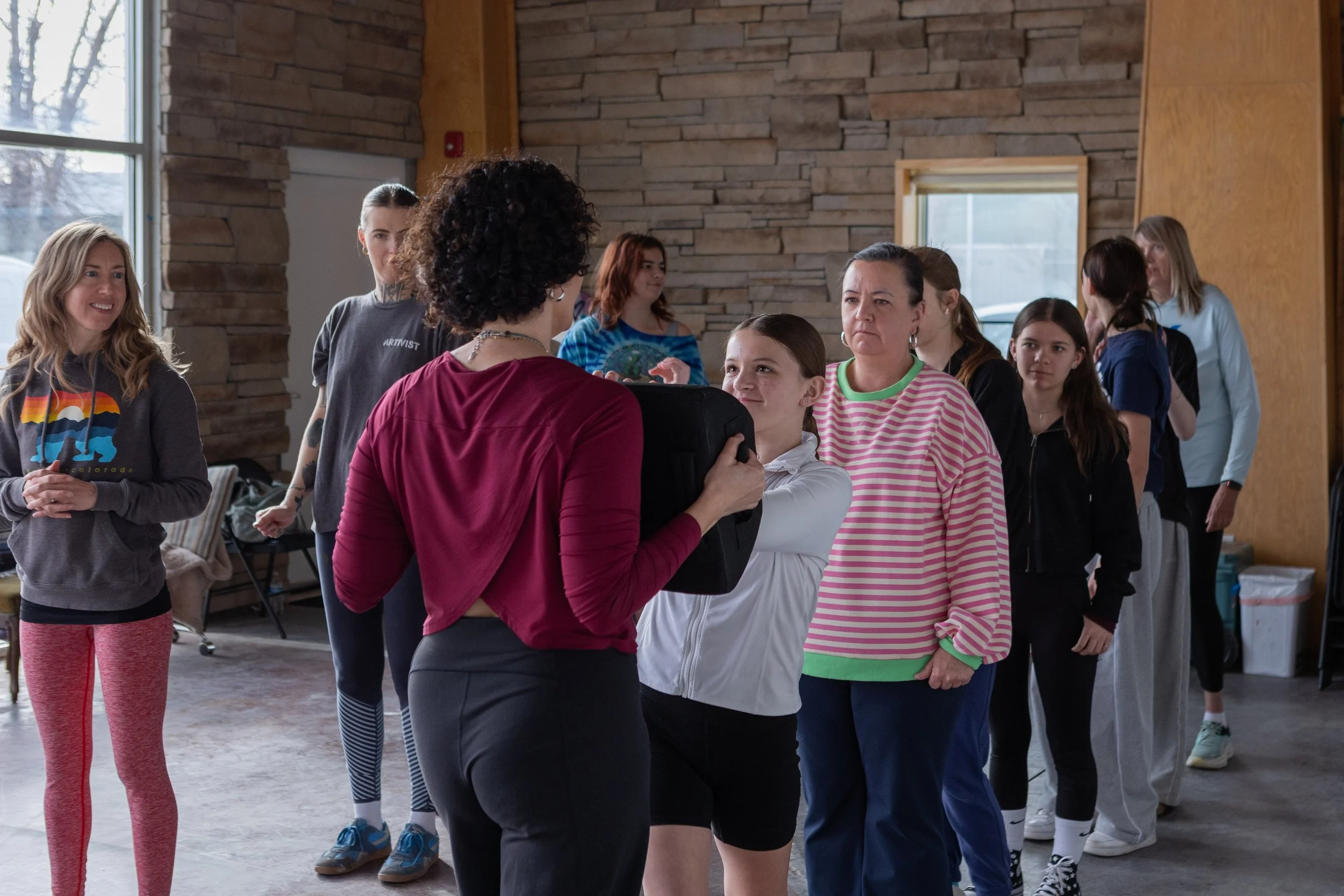 A group of women and girls standing in a line inside a room with stone and wooden walls, as one woman with curly hair and black shorts gives instructions or demonstrating something to a girl in a white jacket. Others are observing and chatting.