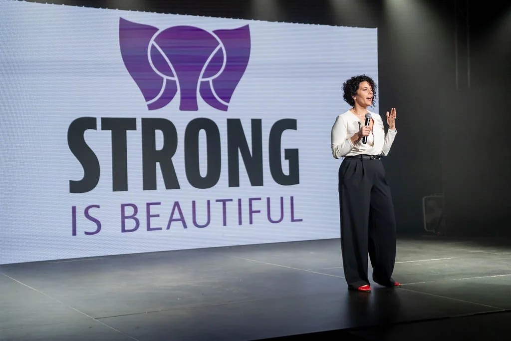 A woman standing on stage holding a microphone, giving a speech or presentation in front of a large screen displaying the logo and slogan 'Strong is Beautiful' with a purple butterfly graphic.