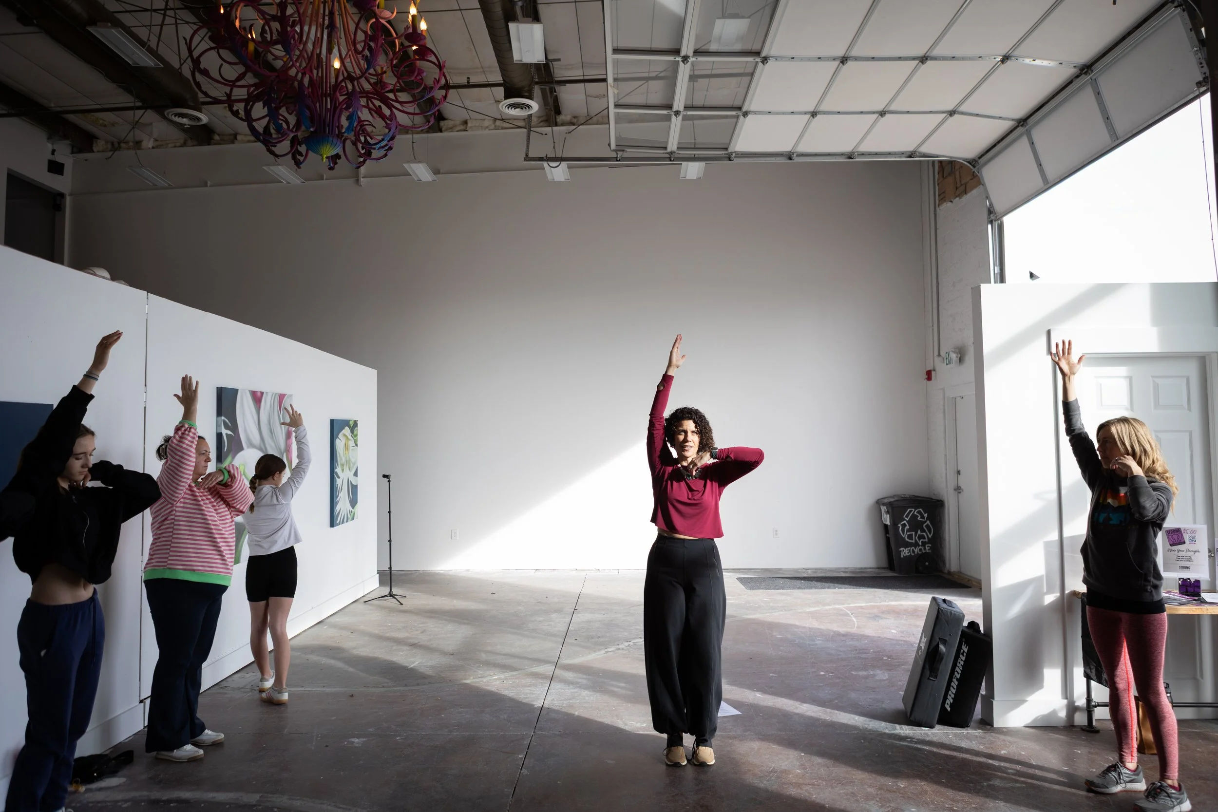 A group of people participating in a fitness or yoga class indoors, following an instructor who is demonstrating a pose. Participants have their hands raised above their heads, with a woman leading in the center. The room has a high ceiling, artwork on the walls, and large open space with natural light coming in from an open garage-style door.