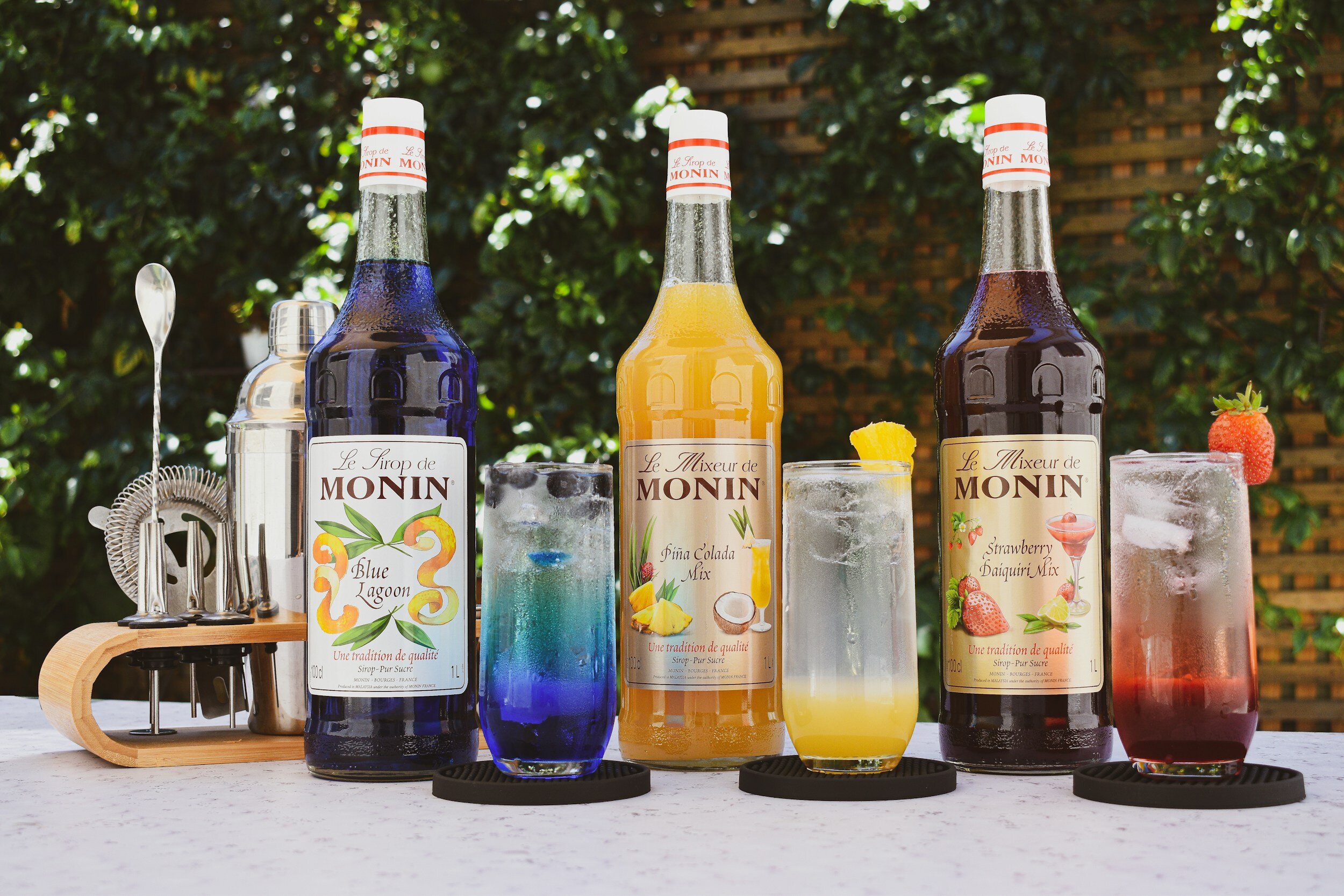 Three bottles of Monin flavored syrup on a table, with three cocktails and cocktail accessories. The syrups are Blue Lagoon, Pine Colada Mix, and Strawberry Daiquiri Mix. The cocktails are colorful, served in tall glasses with ice, garnished with fruit or straw, and placed on black coasters.