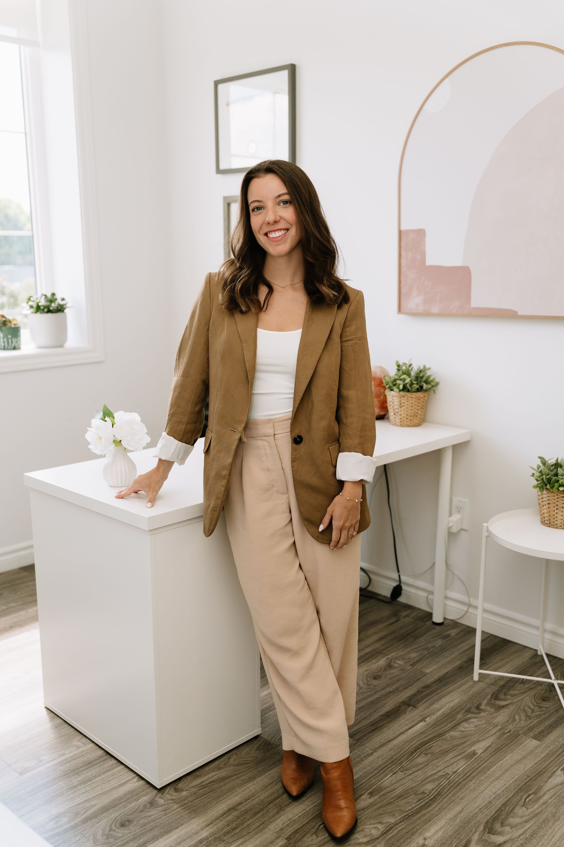 A woman standing in a bright, modern office space, leaning on a white desk with a small flower vase. She is smiling, wearing a tan blazer, beige wide-leg pants, and brown boots. The room has a window with potted plants, framed artwork, and a minimalistic decor.