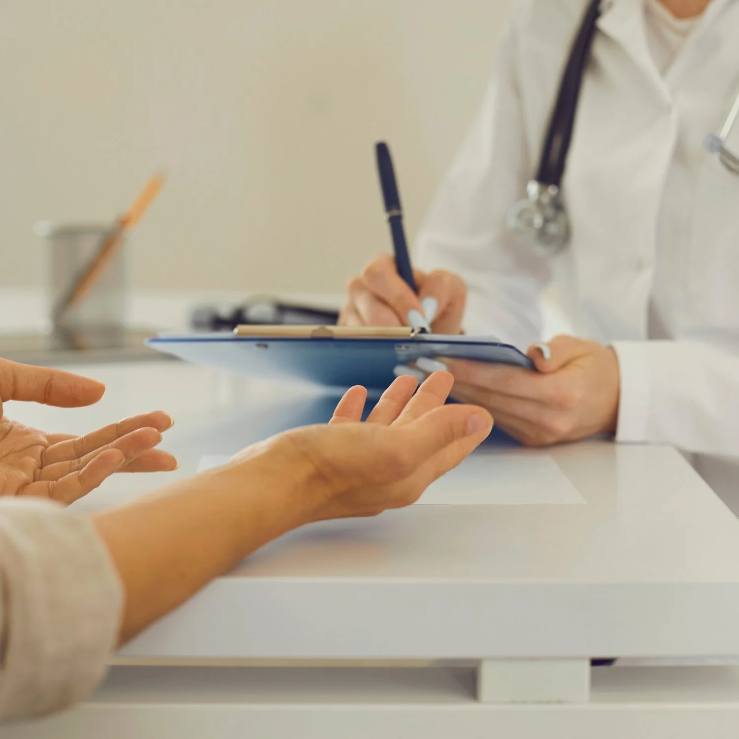 Close-up of a doctor or healthcare professional in a white coat with a stethoscope around their neck writing on a clipboard, while a patient's hand reaches out towards the clipboard during a consultation or medical checkup.