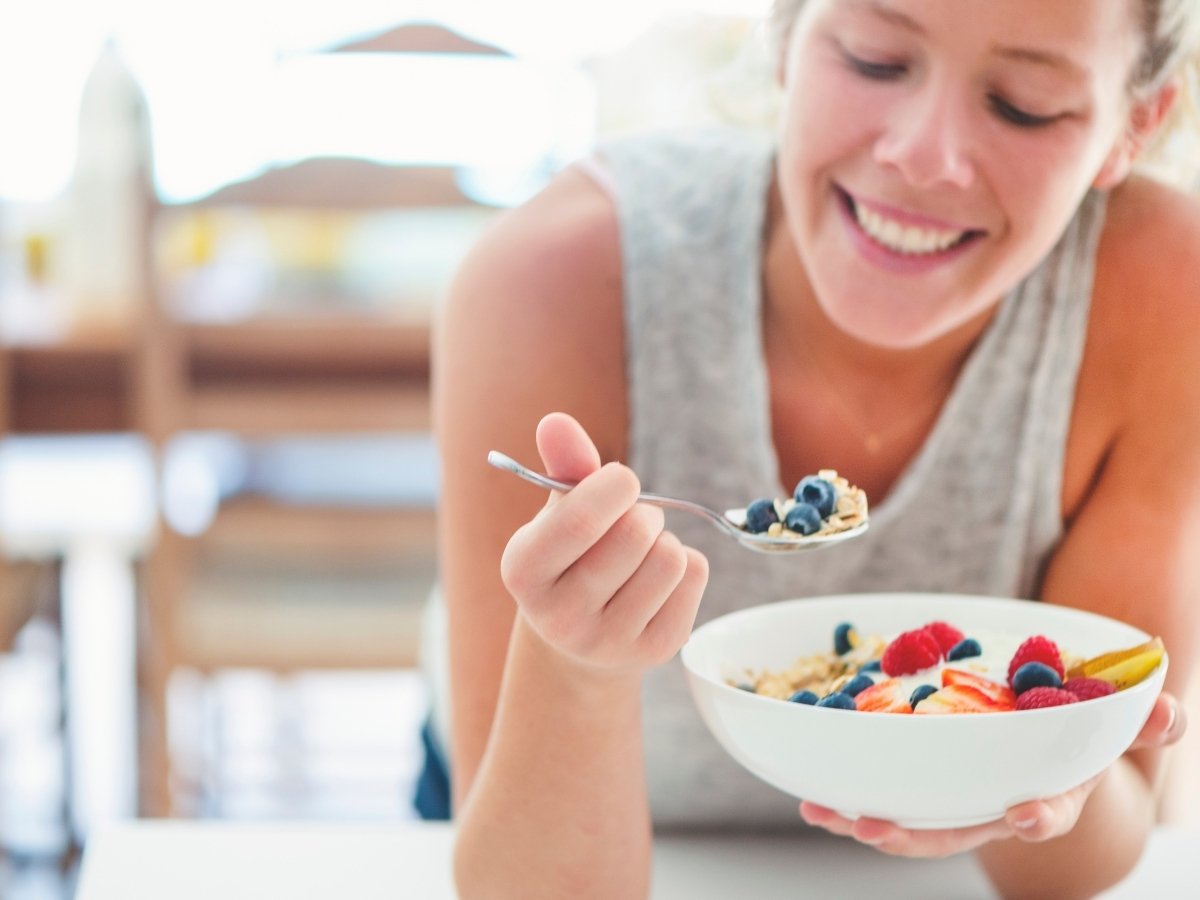 Woman smiling while eating a bowl of cereal with blueberries, raspberries, and sliced strawberries