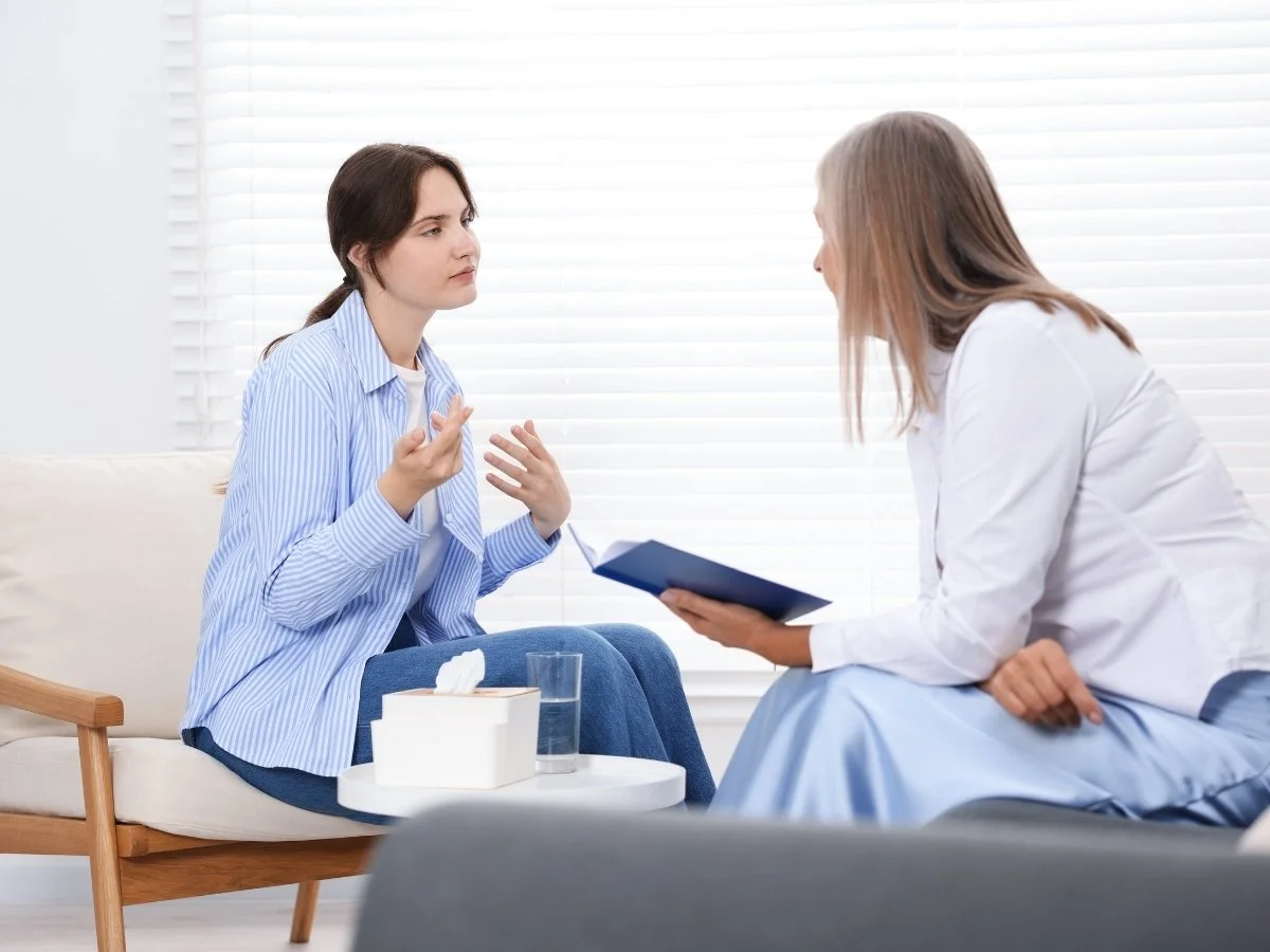 A woman in a blue striped shirt has a serious conversation with a healthcare professional in a white coat, sitting in a medical office with white blinds.