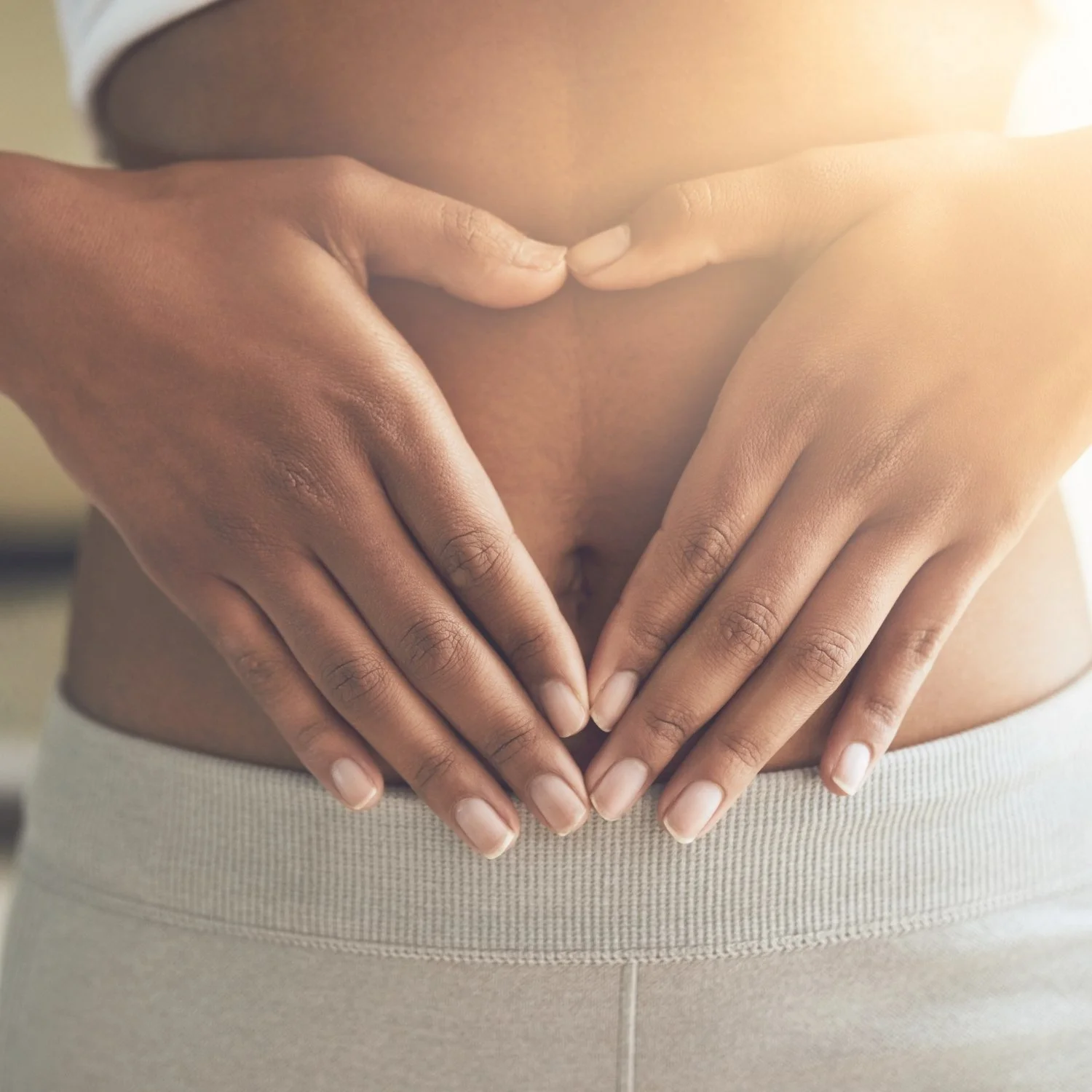 A person with darker skin making a heart shape on their lower stomach using their hands.