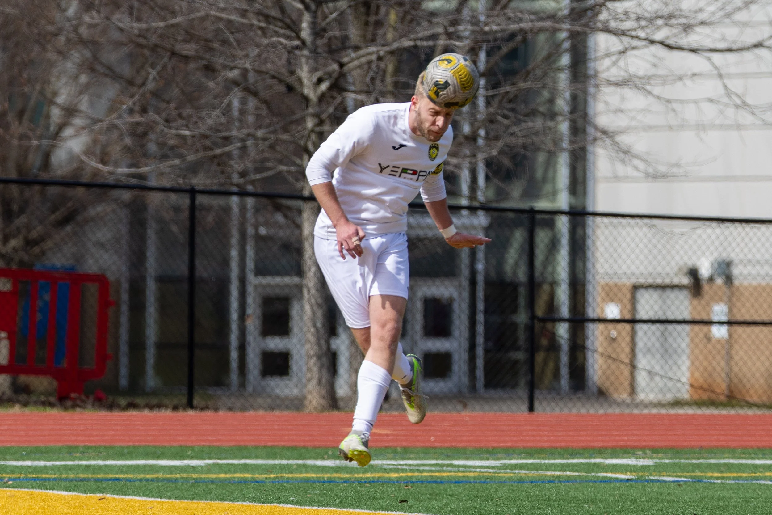 A soccer player on a field, jumping for the ball, wearing a white jersey and shorts, with a colorful ball on his head.