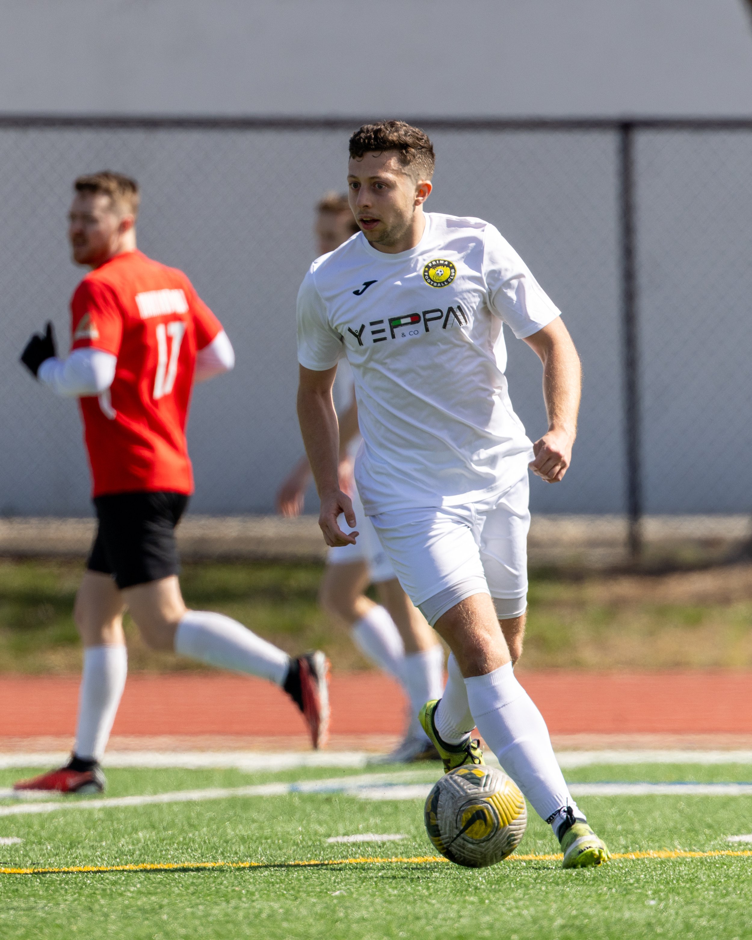 A soccer player in a white uniform dribbling a ball on the field, with other players in the background.