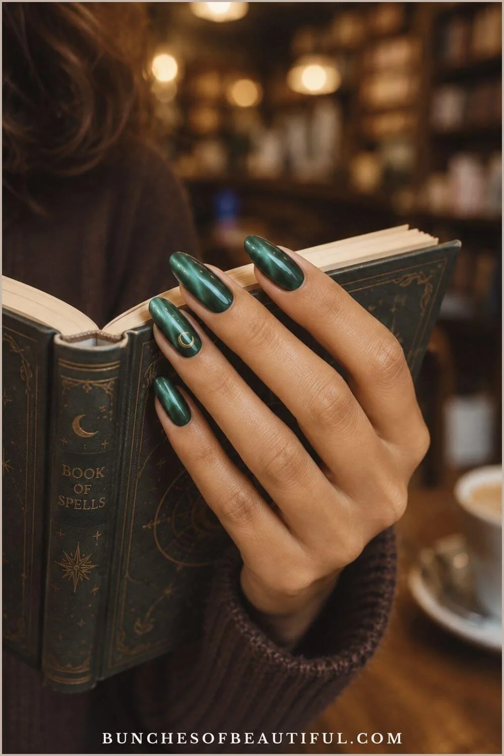 Shimmering emerald cat-eye nails with a small gold crescent moon while holding an open spell book.