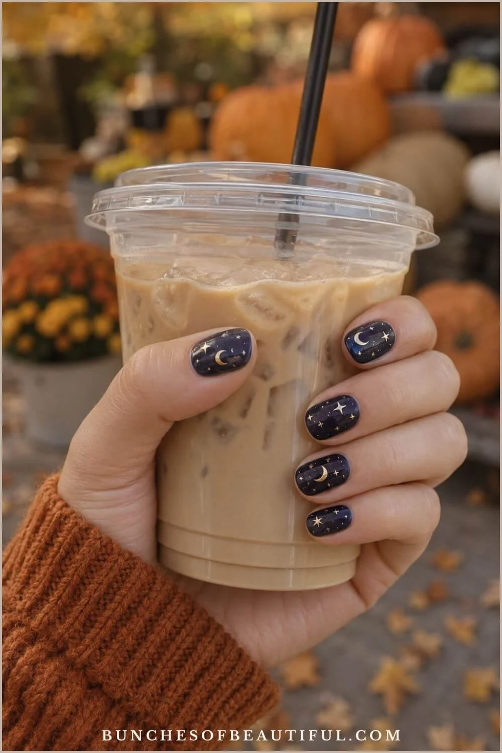 Short navy blue nails decorated with gold crescent moons and stars while holding an iced coffee at a pumpkin patch.