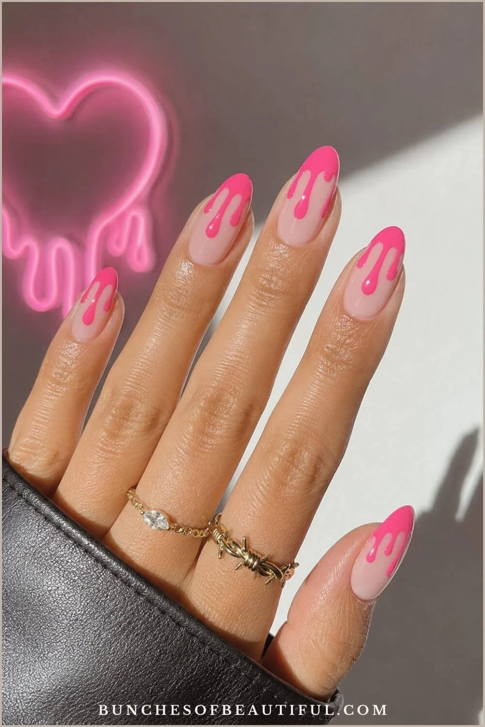 Nude almond nails with bright hot-pink polish dripping from the tips, posed beside a glowing pink neon heart.