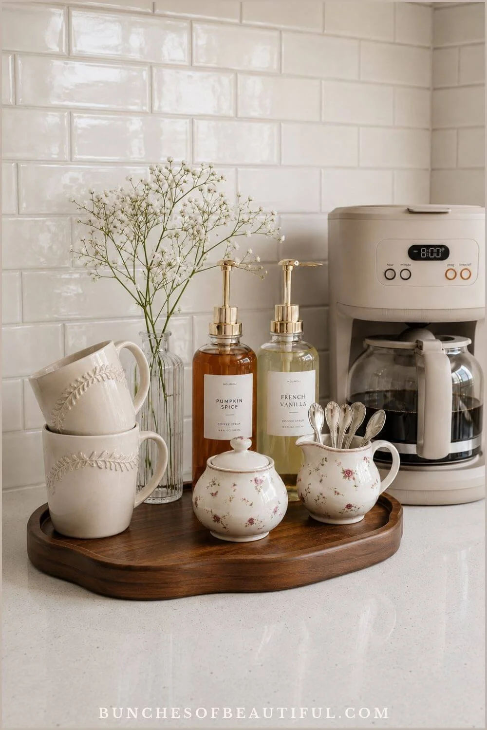 Cottage kitchen counter with a drip coffee maker, wood tray, floral pottery, stacked cream mugs, syrup bottles, and spoons.