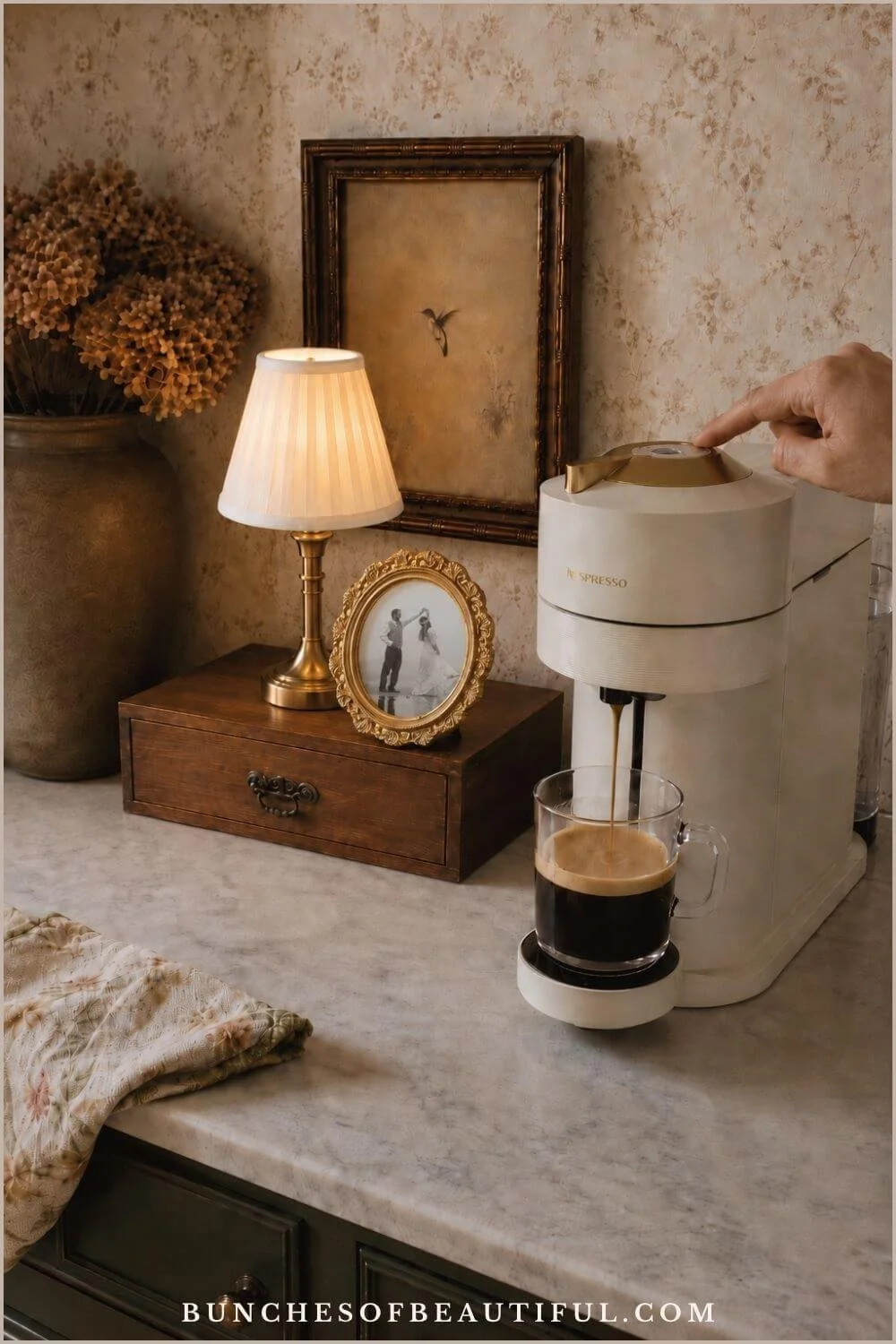 Vintage coffee corner with a compact cream brewer, small lamp, floral wallpaper, framed artwork, and a dark wood storage box.