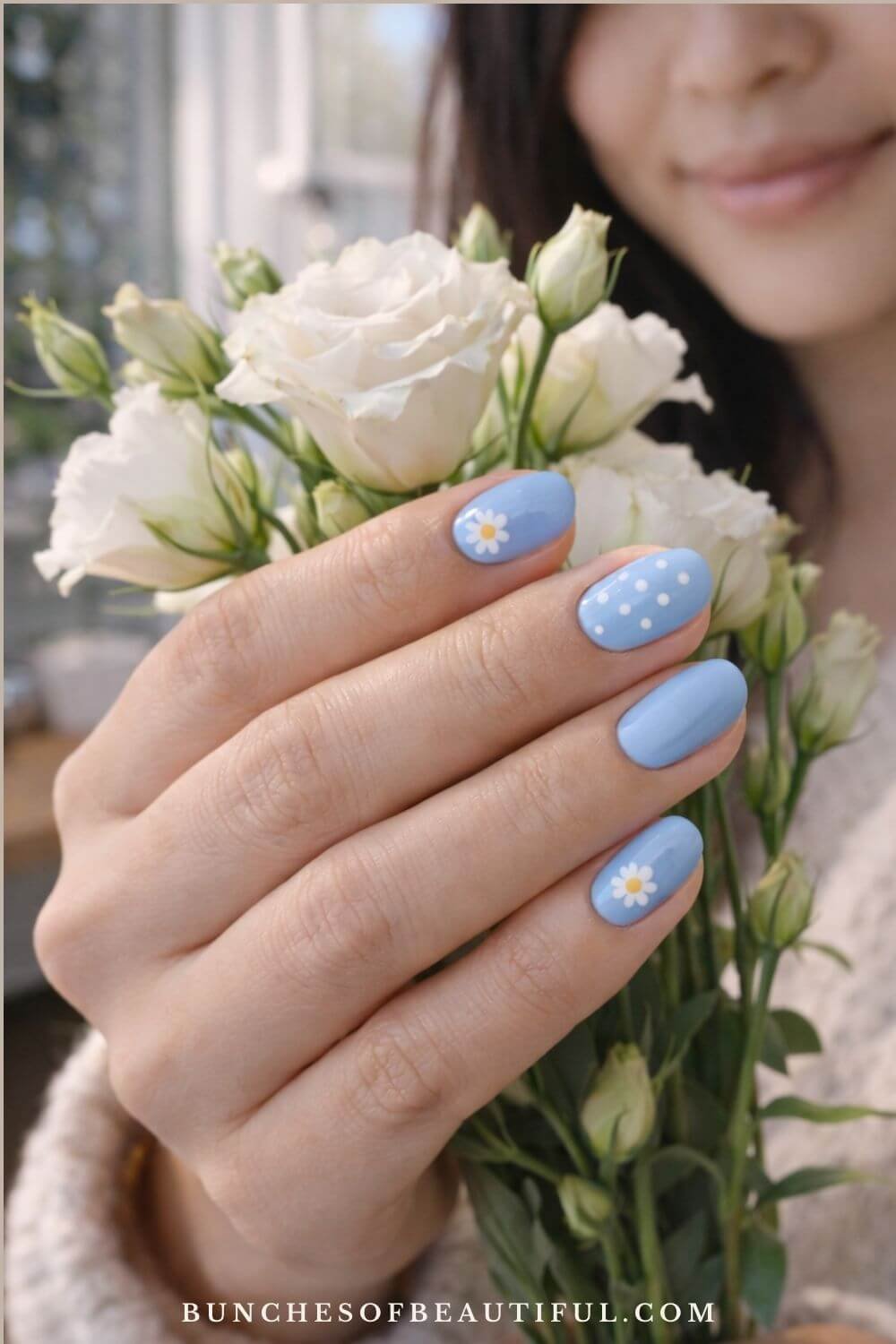 Pastel blue spring nails with daisy flower nail art and white polka dots photographed while holding a bouquet of white flowers.