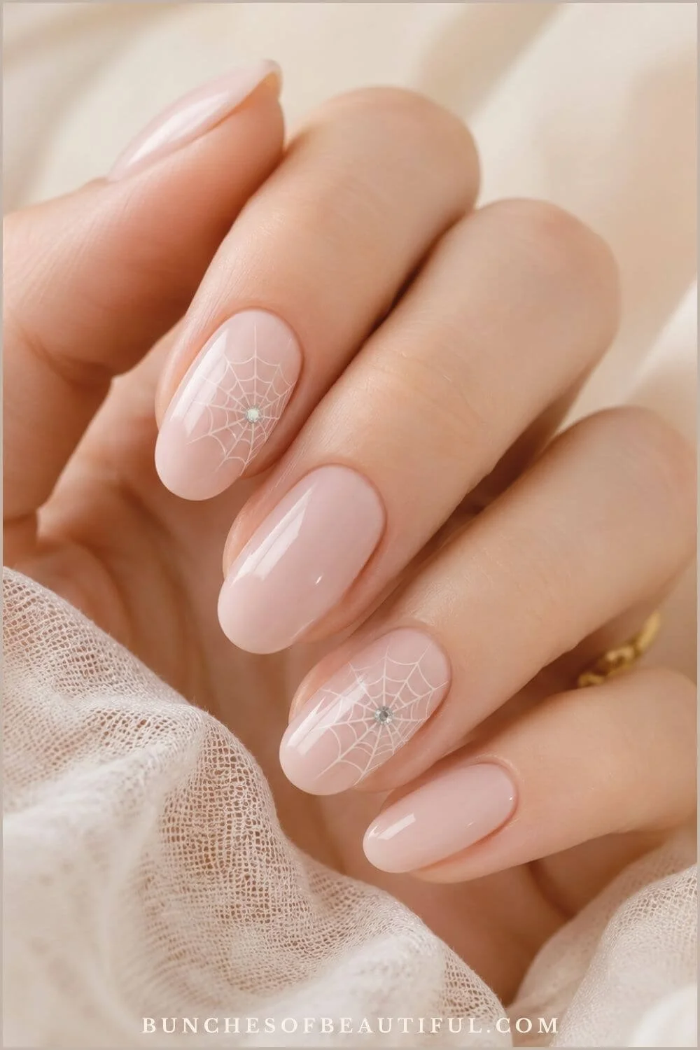 Glossy blush-pink oval nails with delicate white spiderweb accents and tiny rhinestone centers against soft ivory fabric.
