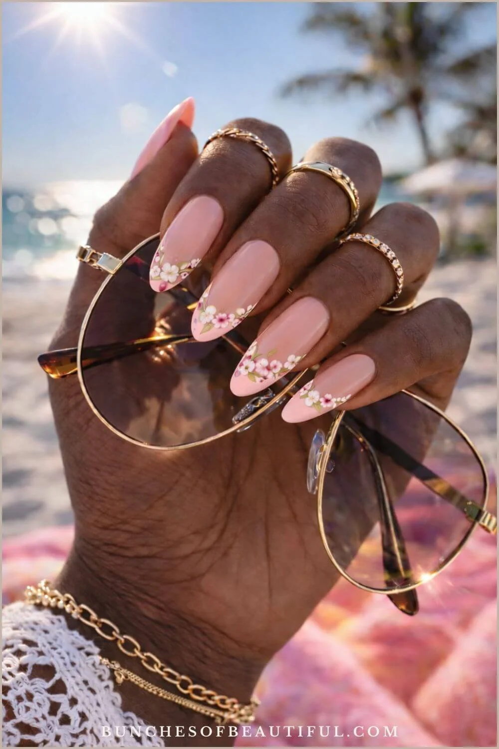 Peach nude spring nails with delicate floral tip design photographed on a beach with sunglasses and gold jewelry.