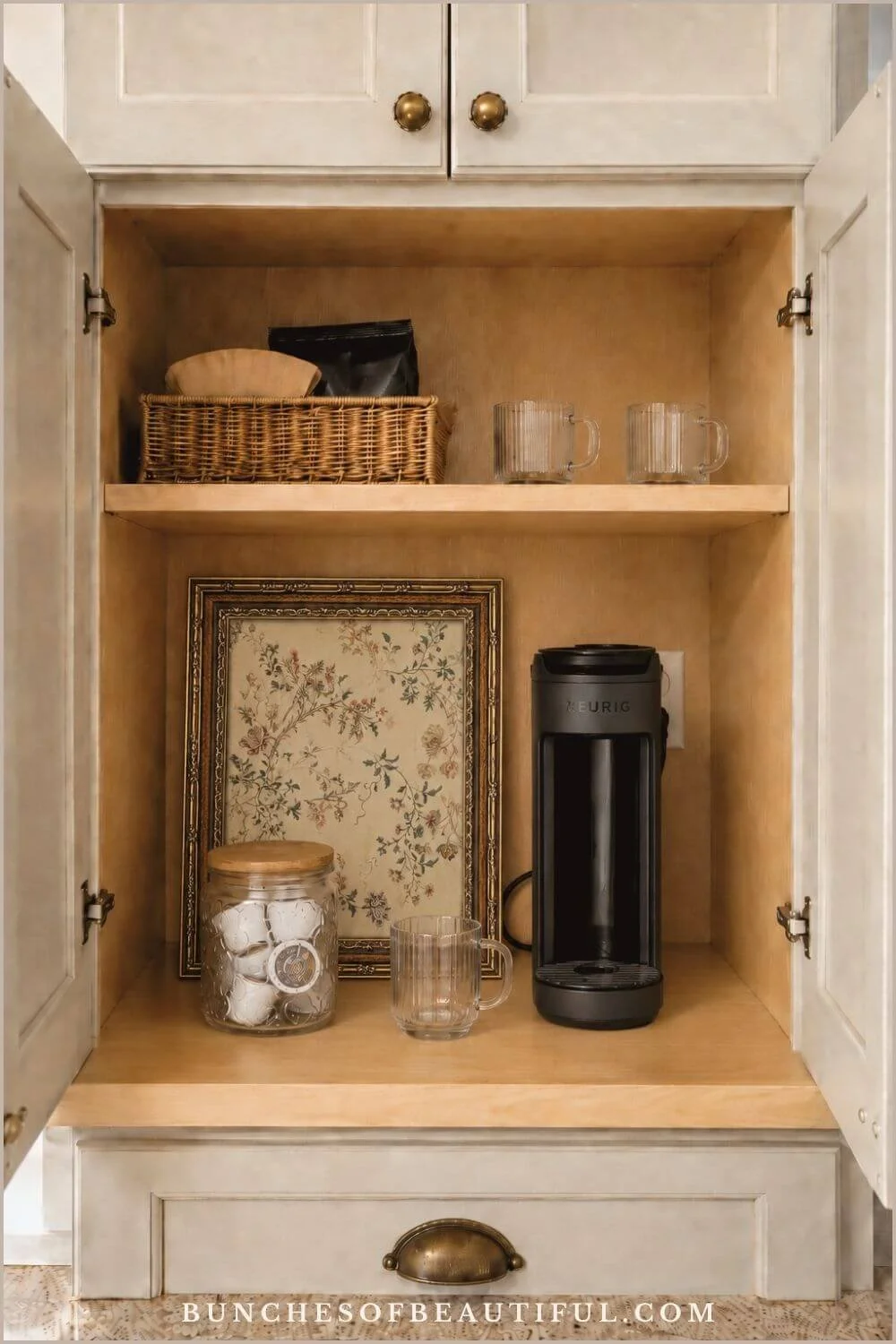 Open wood cabinet containing a slim single serve coffee maker, glass mugs, clear pod canister, storage basket, and botanical artwork.