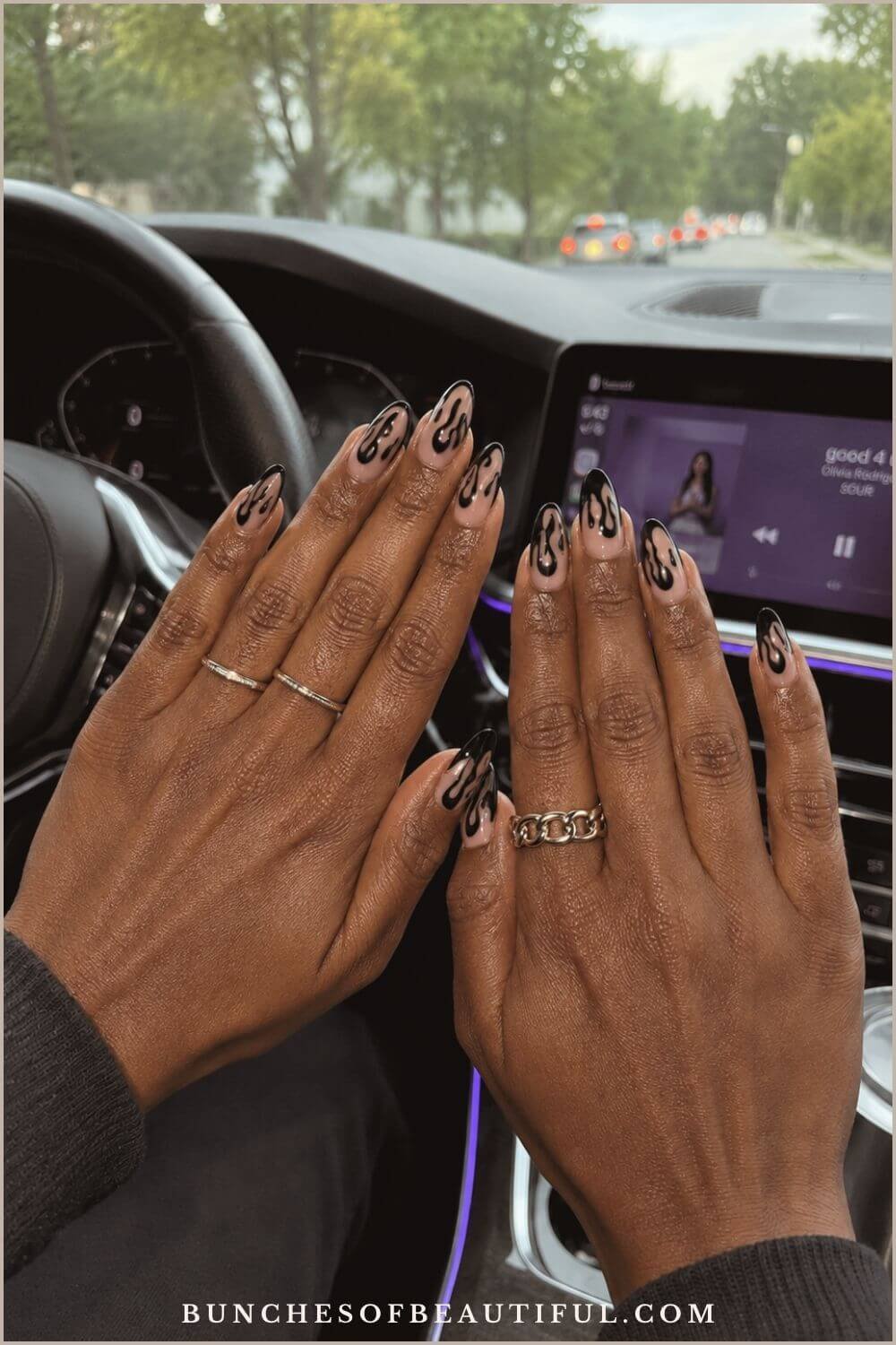 Two hands with nude almond nails and glossy black flame tips posed inside a modern car.
