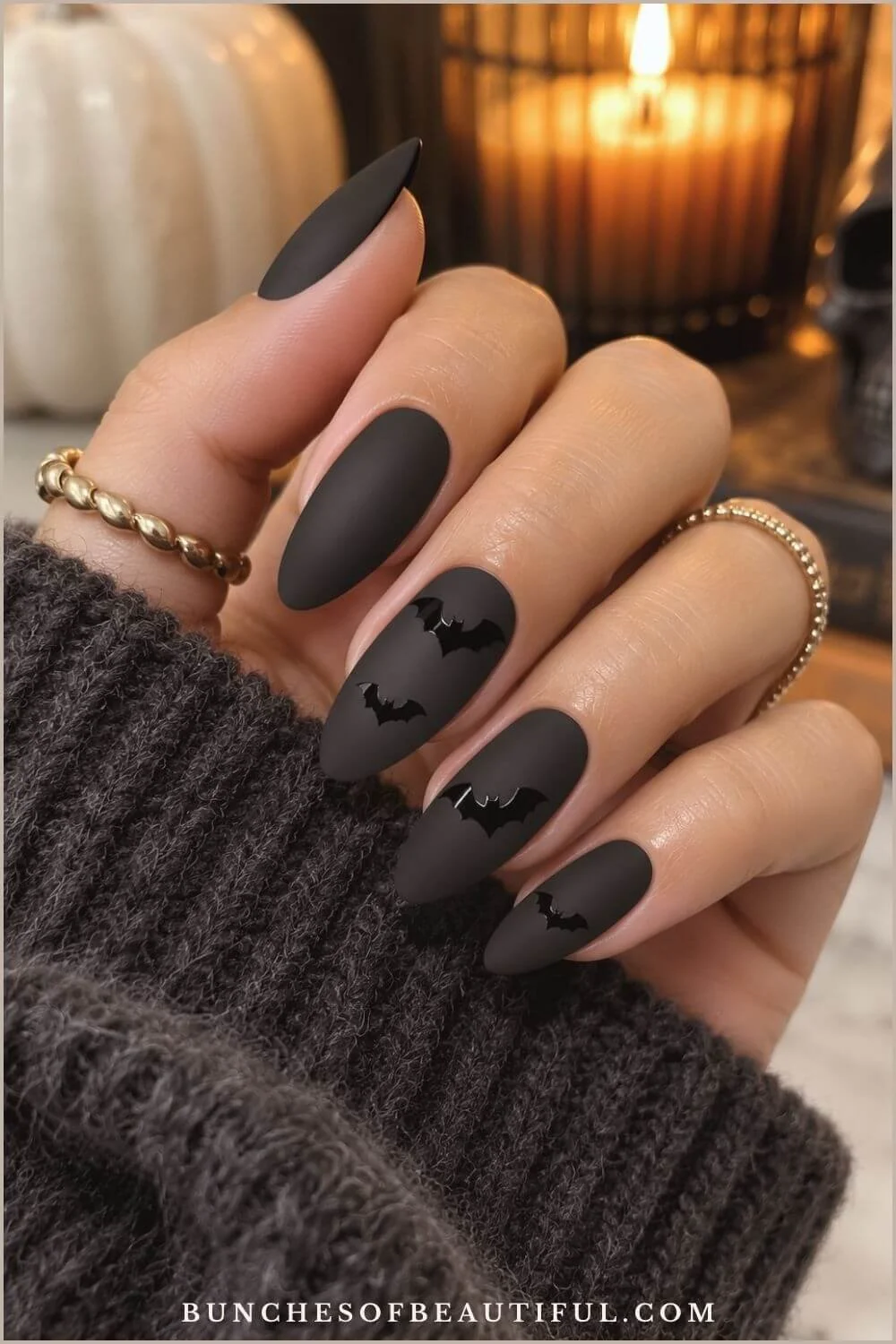 Matte black almond nails decorated with glossy black bat silhouettes in a cozy Halloween setting.